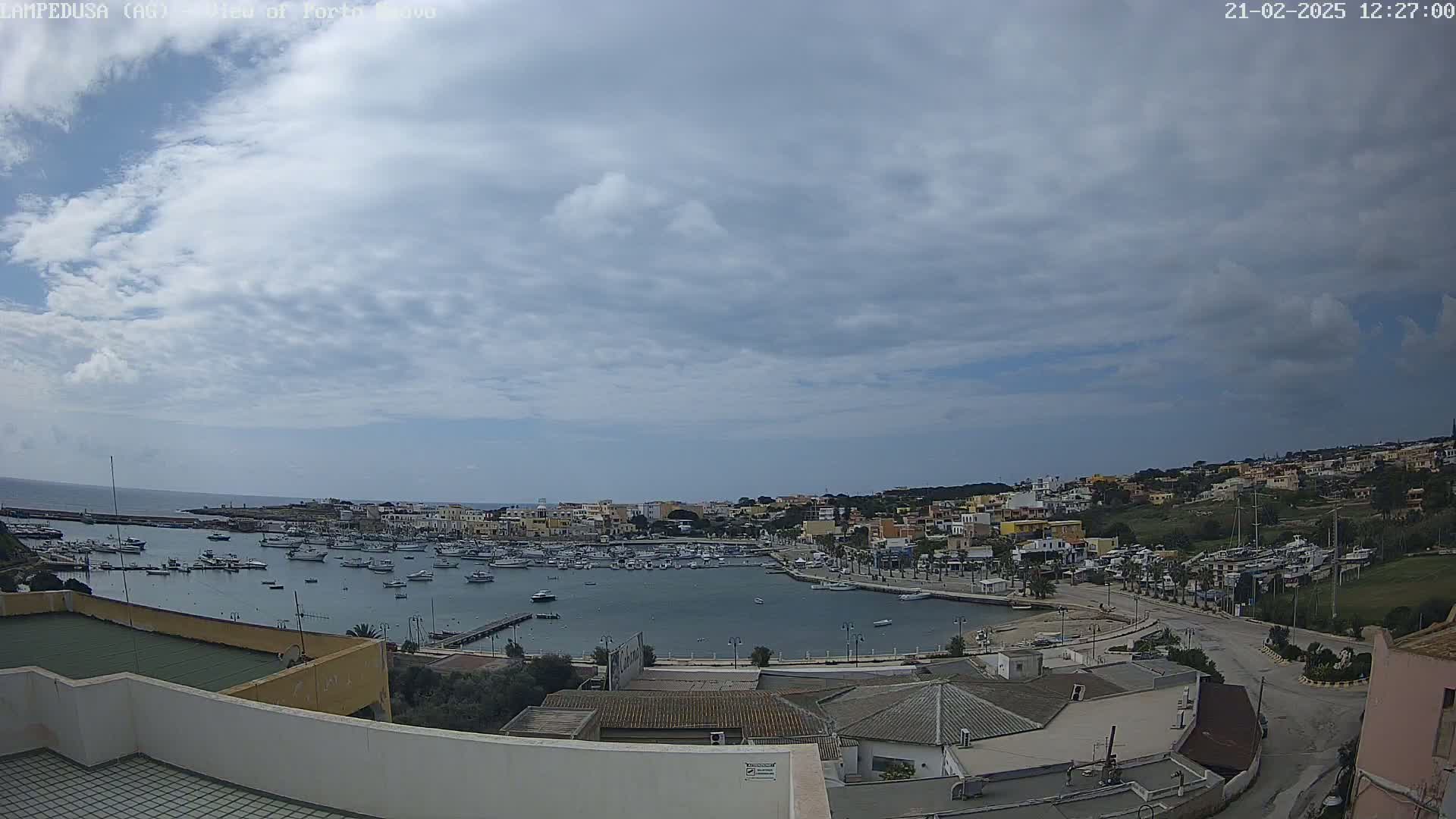 An overcast sky overlooks a harbor filled with boats and a town of low-rise buildings nestled on a hillside.