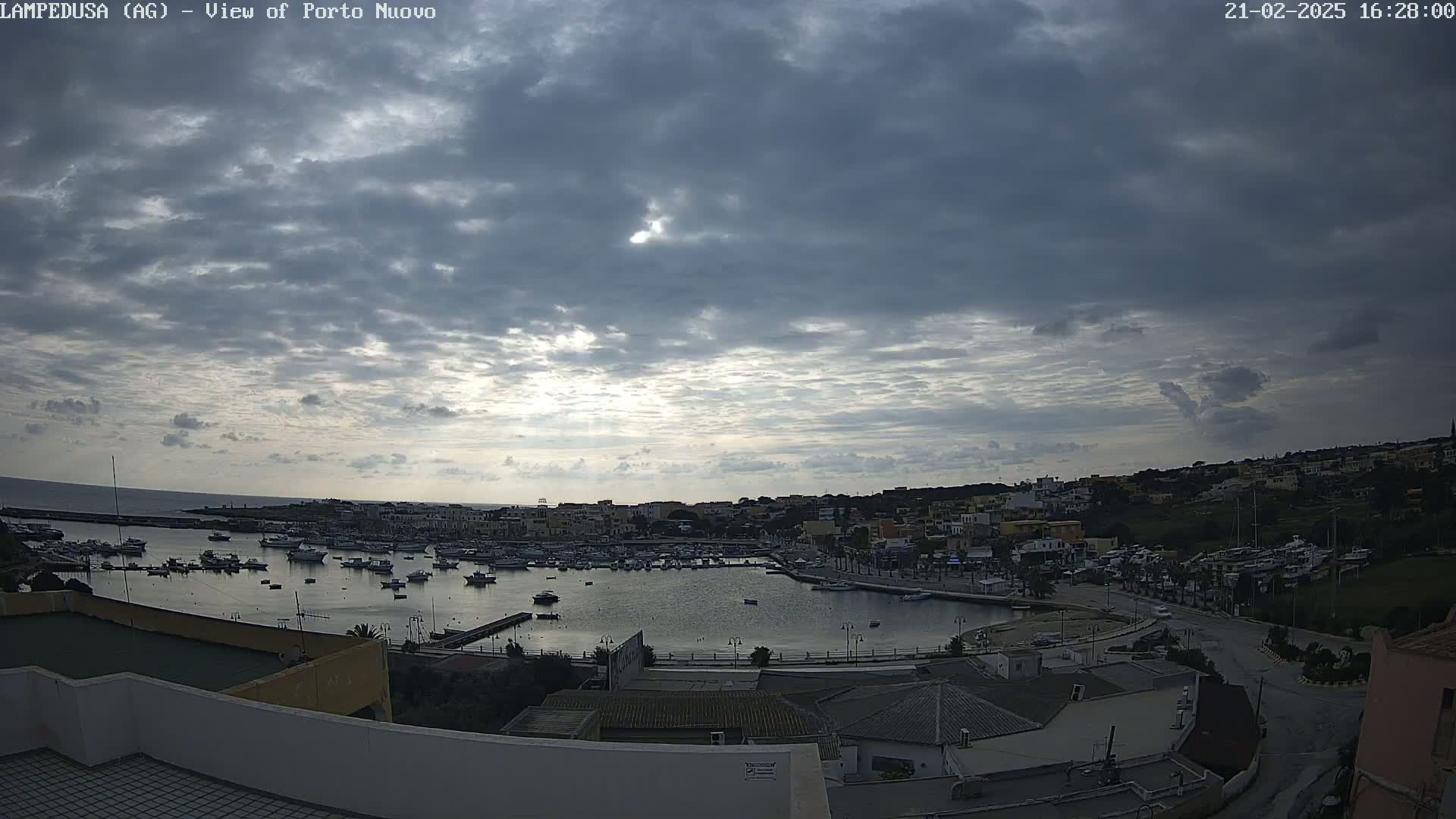 A harbor filled with boats is situated below a mostly cloudy sky with some sunlight breaking through.