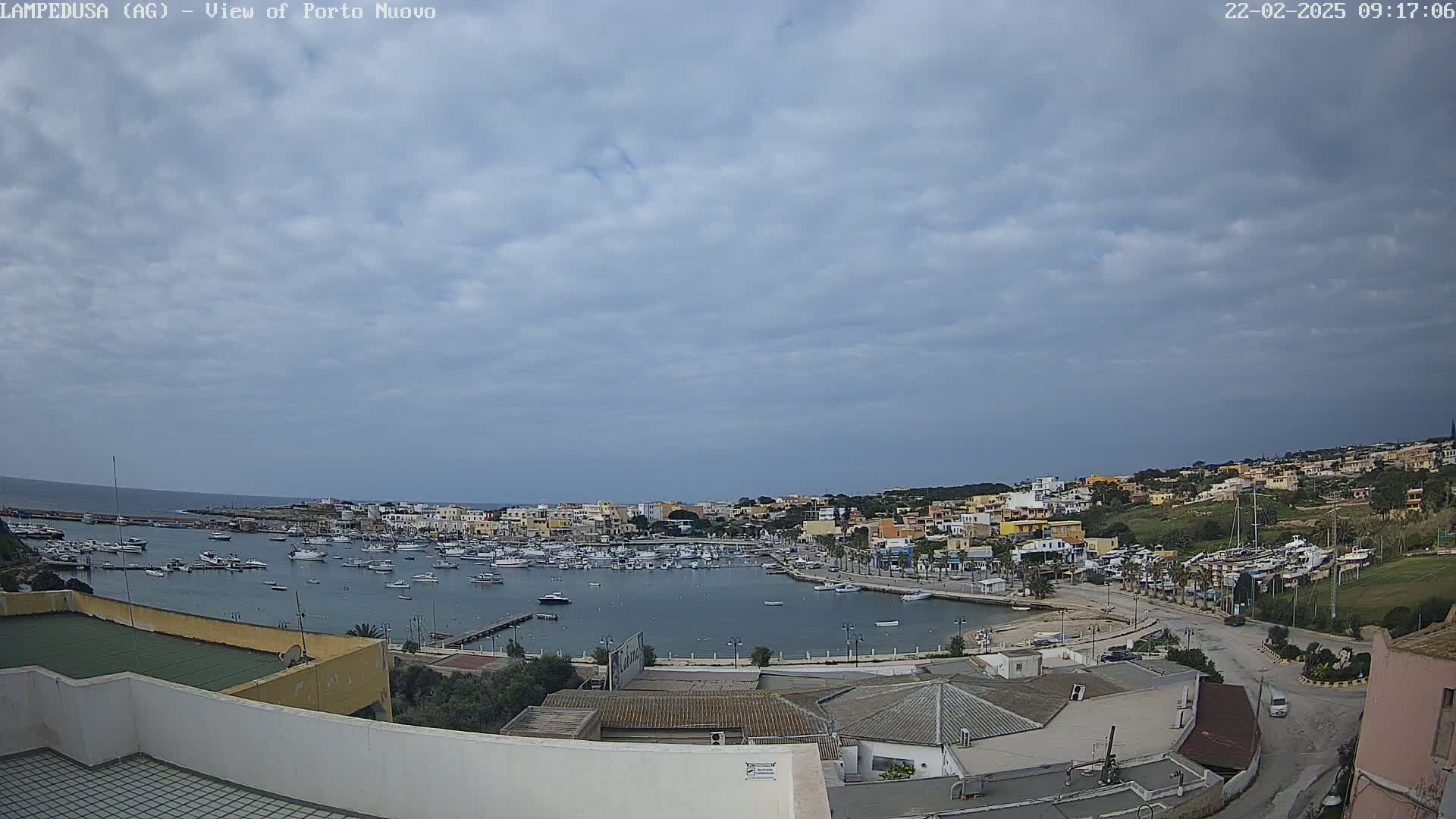 A harbor filled with boats is nestled in a town of low-rise buildings under a mostly cloudy sky.