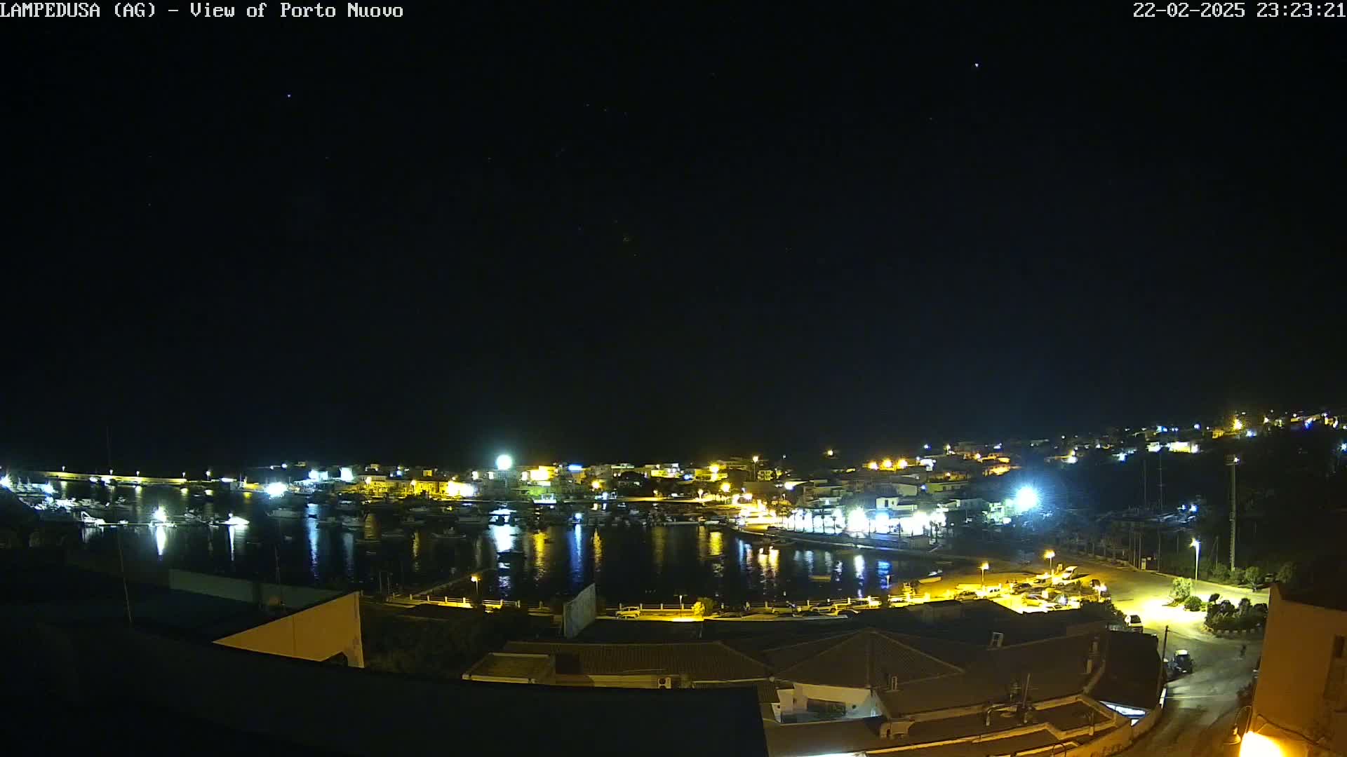 A nighttime, aerial view of a harbor town with boats in the water and buildings illuminated along the shoreline under a clear, dark sky.