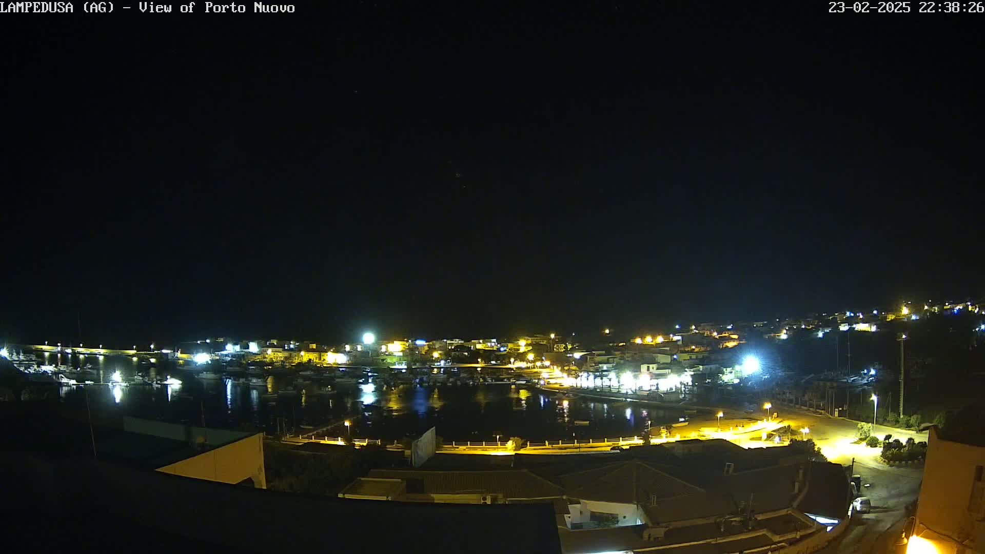 A nighttime, aerial view of a harbor town with boats docked, illuminated buildings, and clear skies.