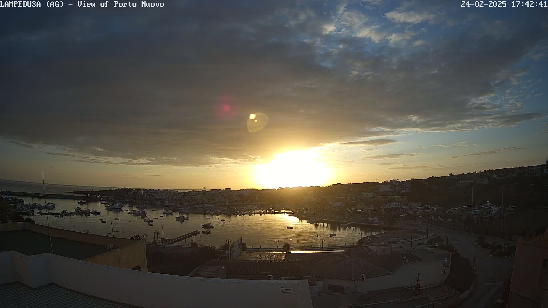 A sunset over a harbor filled with boats and a town, under a partly cloudy sky.
