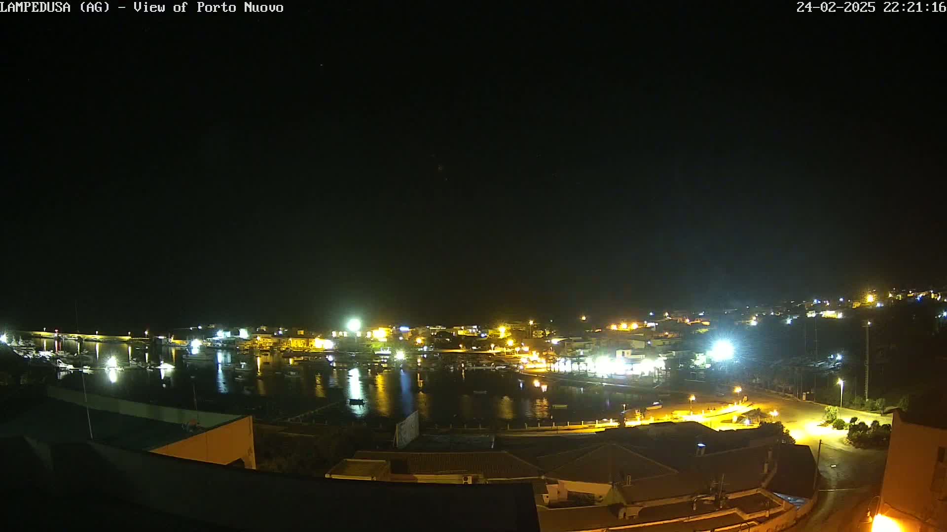 A nighttime view of a harbor town with its lights reflecting on the calm water under a clear, dark sky.