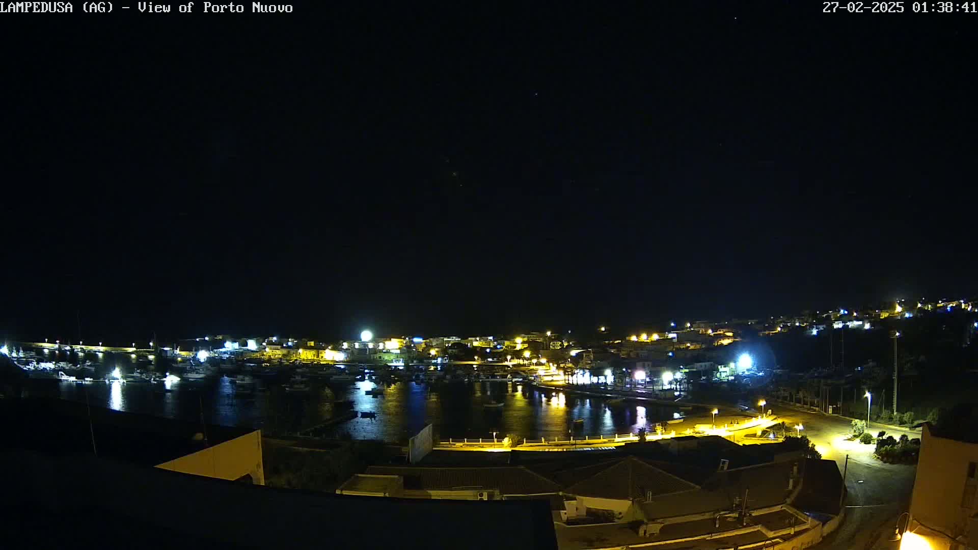 A nighttime view of a harbor town with boats and buildings illuminated, under a clear, dark sky.