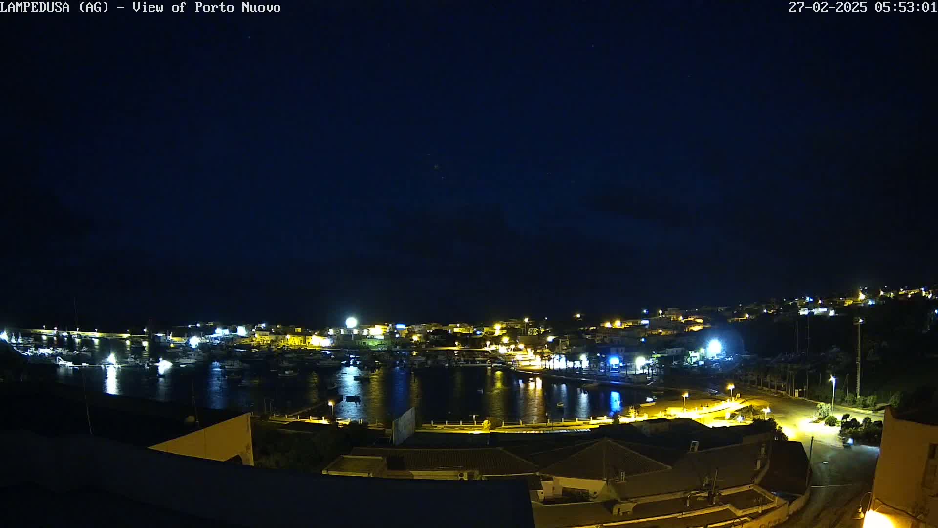 A nighttime, high-angle view of a harbor town with boats in the water and buildings illuminated by lights under a dark, clear sky.