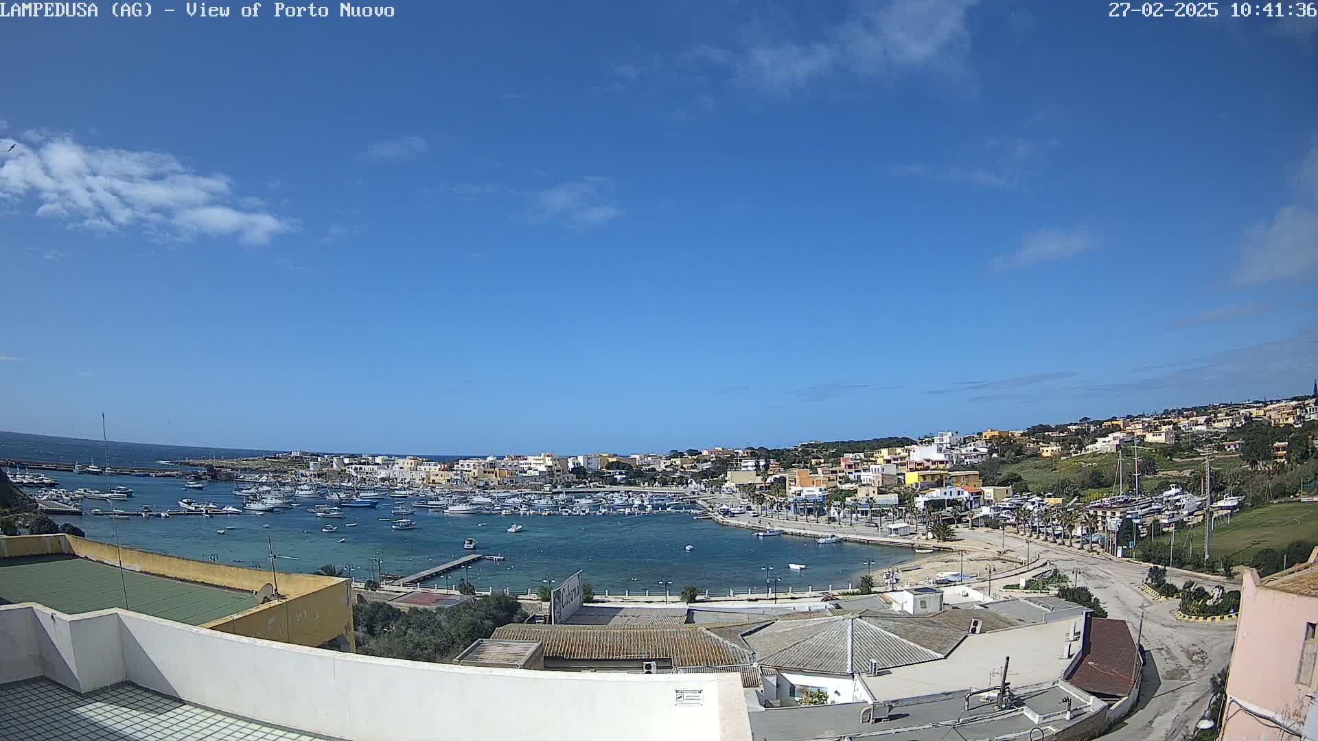 A mostly sunny day overlooking a harbor filled with boats and a town of low buildings on a hillside.