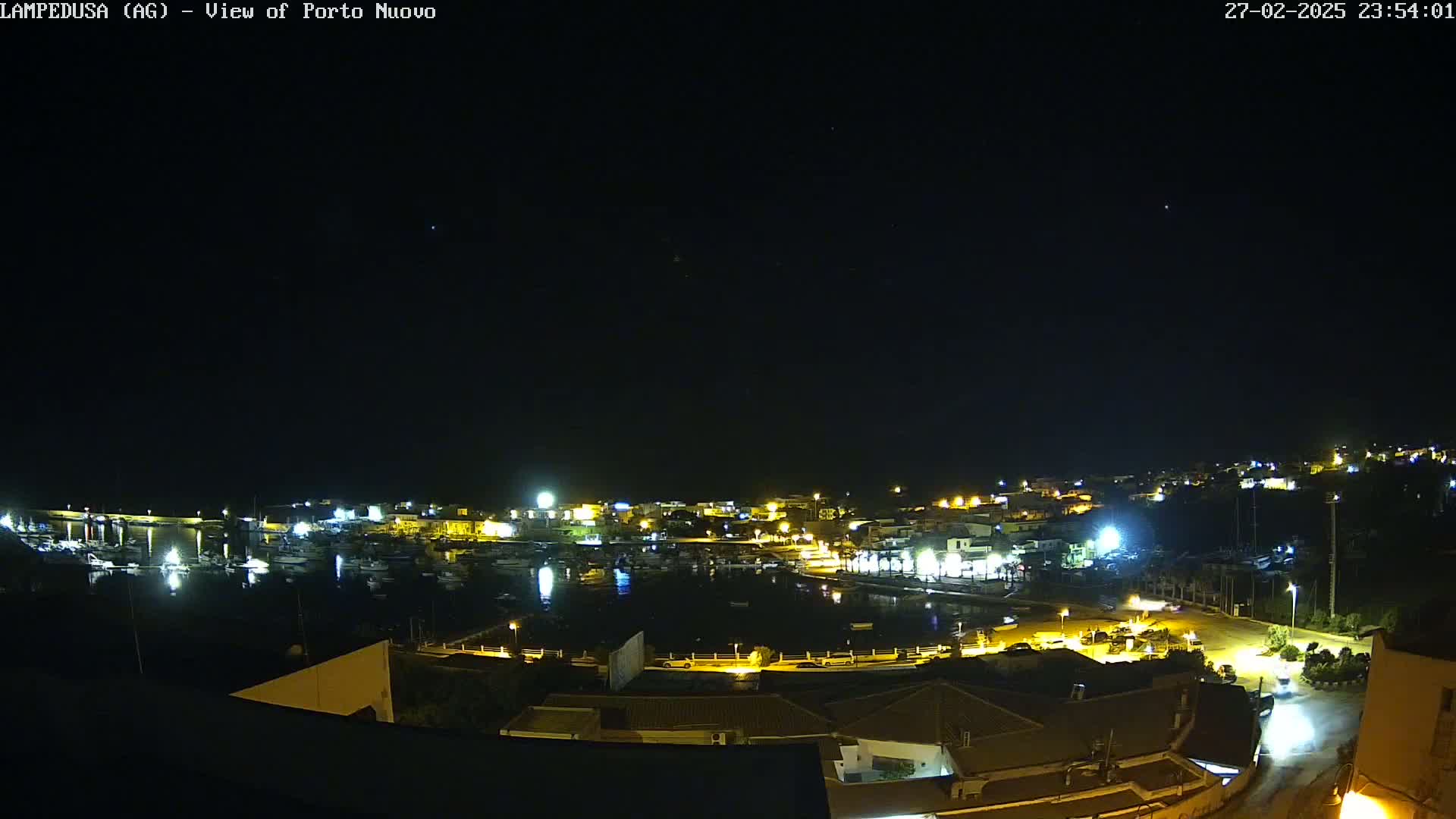 A nighttime view of a harbor town with boats in the water and buildings illuminated by lights under a clear, starry sky.