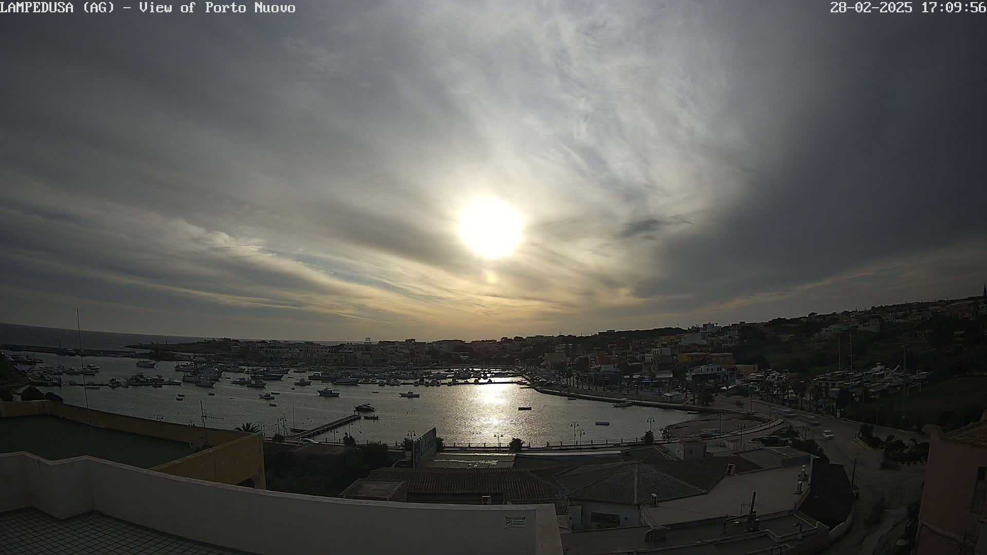 A harbor filled with boats is seen from an elevated viewpoint under a partly cloudy sky, with the sun shining brightly.