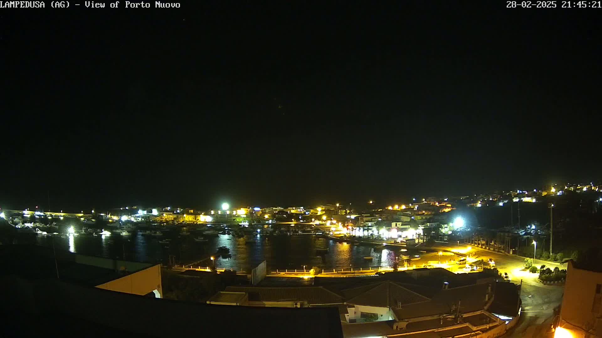 A nighttime, aerial view of a harbor town with boats in the water and buildings lit up along the shoreline under a clear, dark sky.