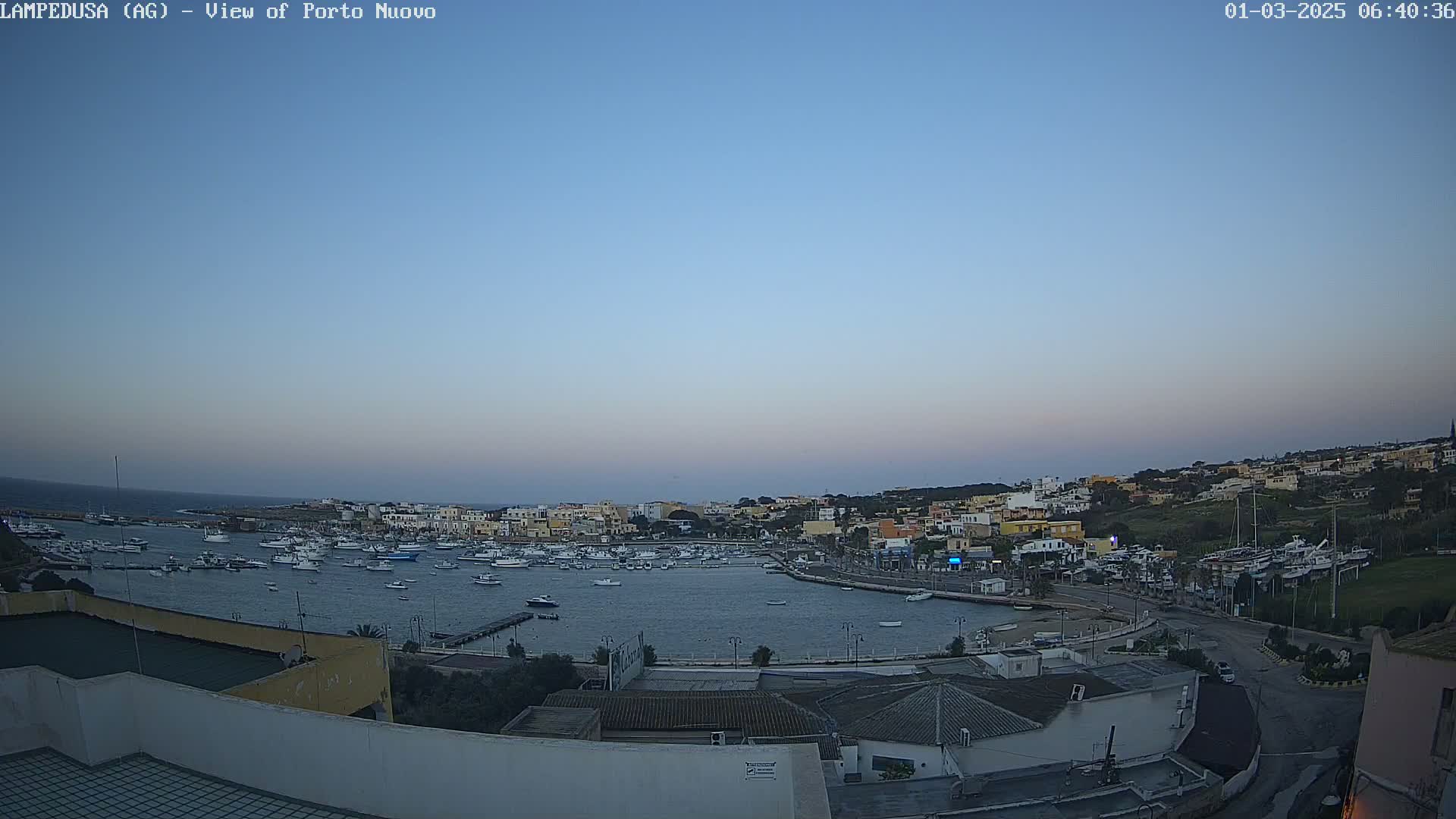 A harbor filled with numerous boats is nestled within a town situated on a hill overlooking the calm, twilight sea.