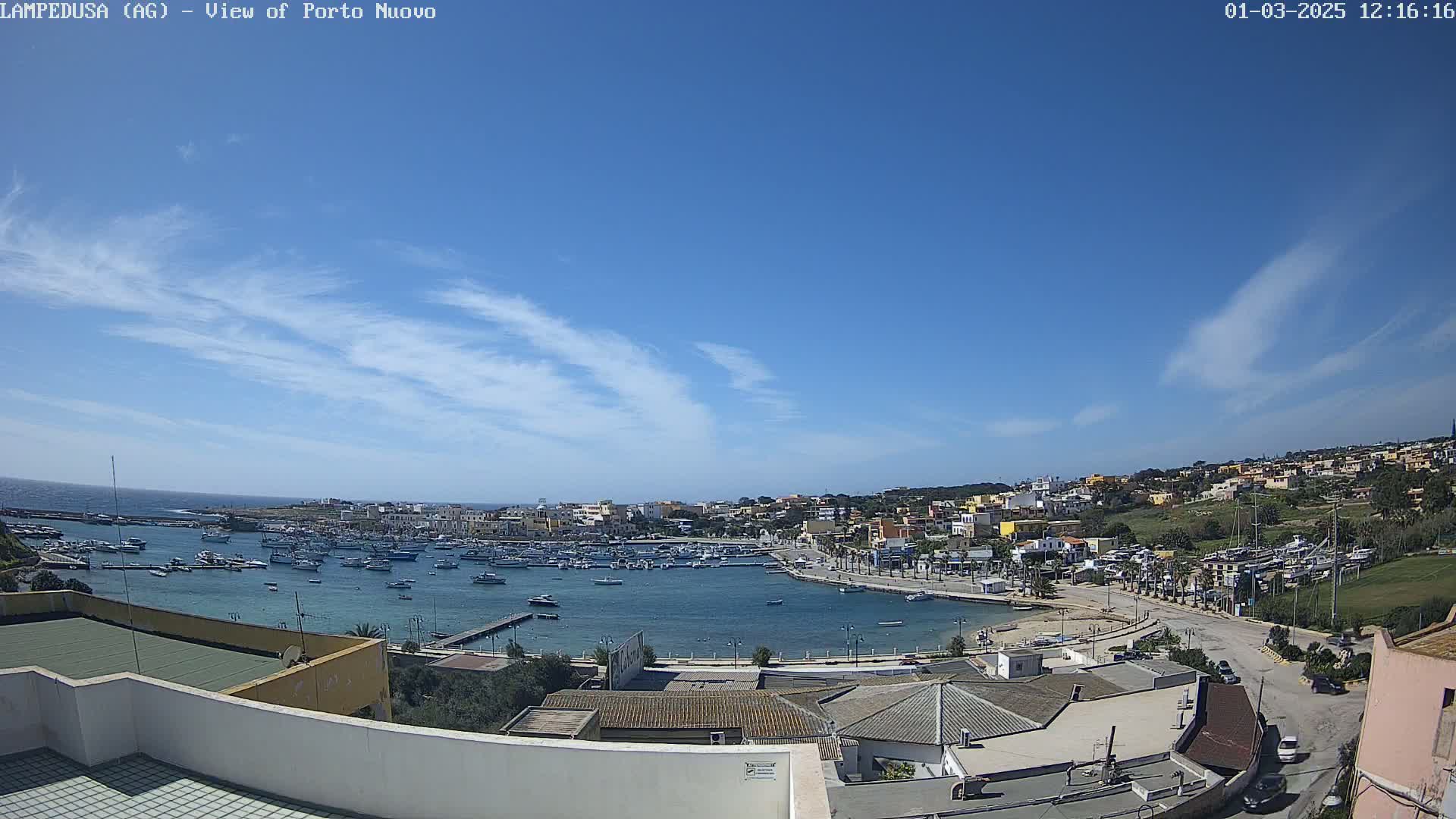 A harbor filled with boats is surrounded by a town of low-lying buildings under a mostly clear blue sky with a few wispy clouds.