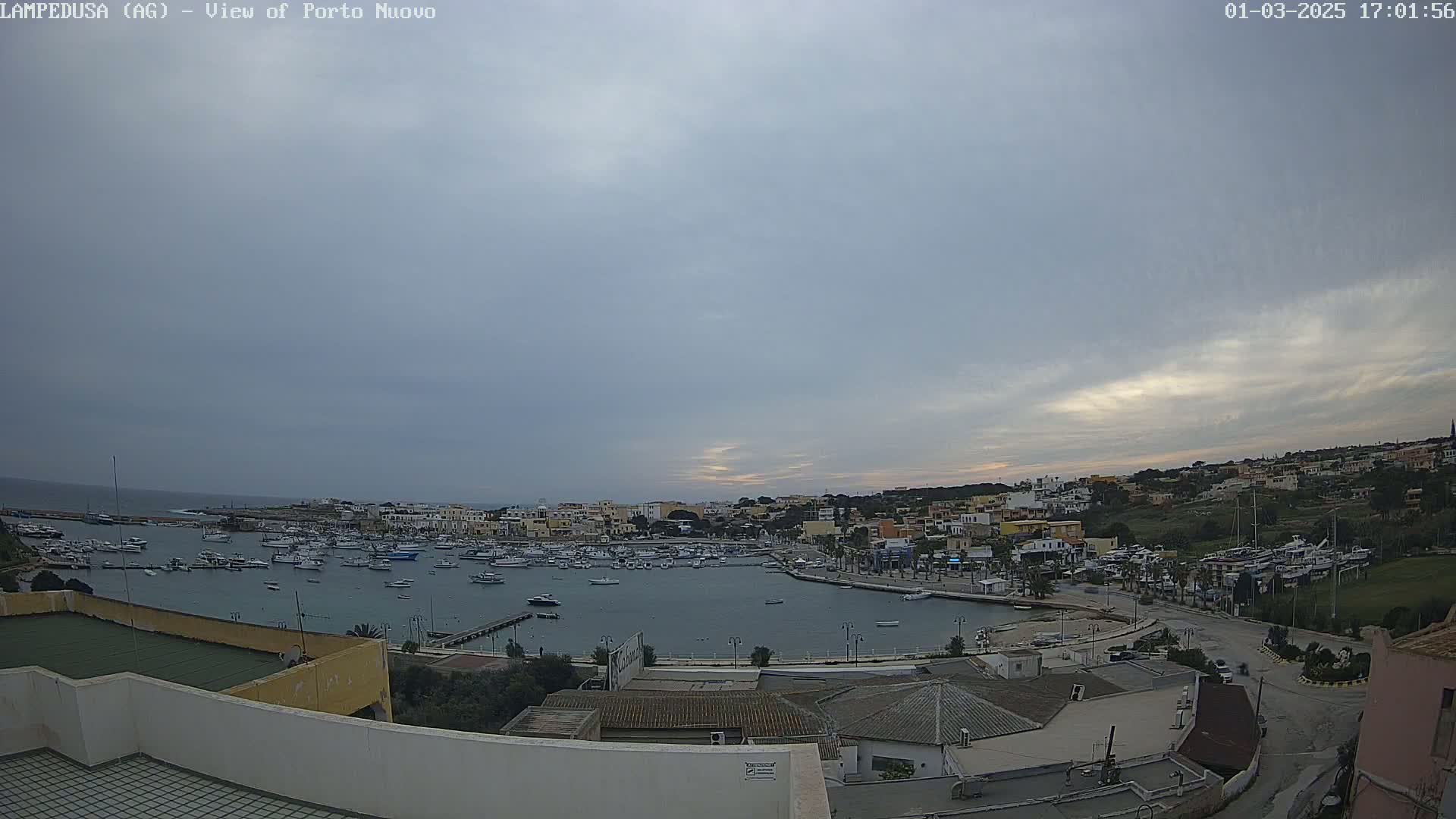 A harbor filled with boats is nestled in a coastal town under a mostly cloudy sky.