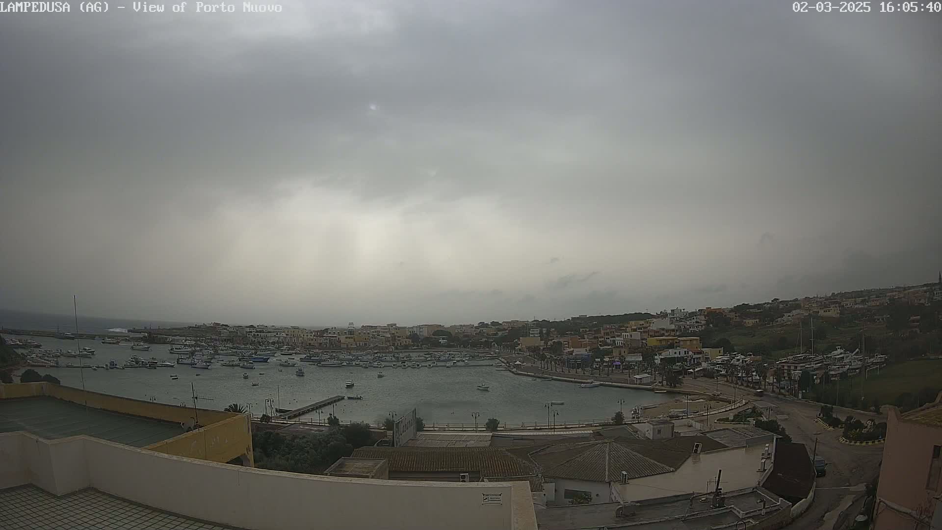 An overcast sky hangs over a harbor filled with numerous boats, a town with low buildings, and roads.