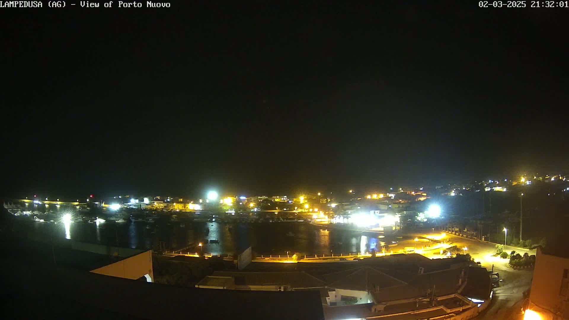A nighttime, aerial view of a harbor town with many lights illuminating the buildings, streets, and boats in the water under a clear, dark sky.
