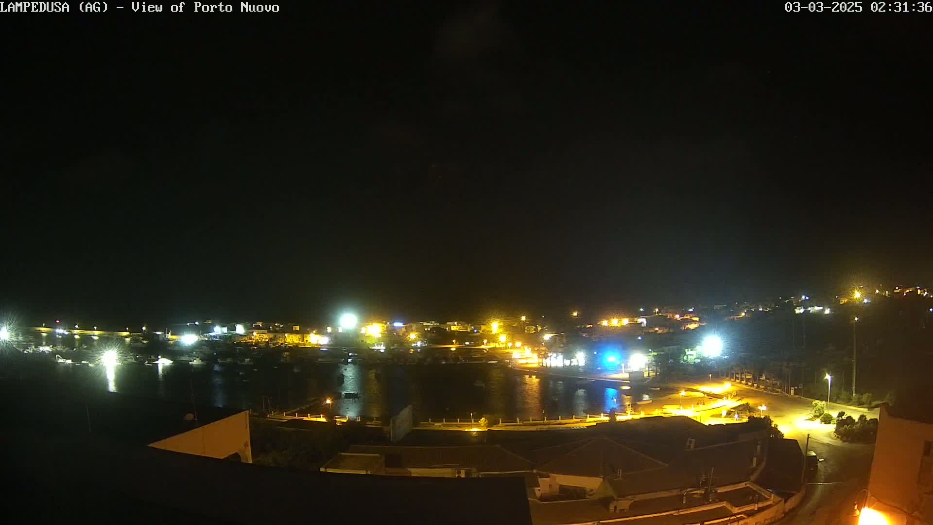 A nighttime view of a harbor town with many lights illuminating the buildings and water, under a clear, dark sky.
