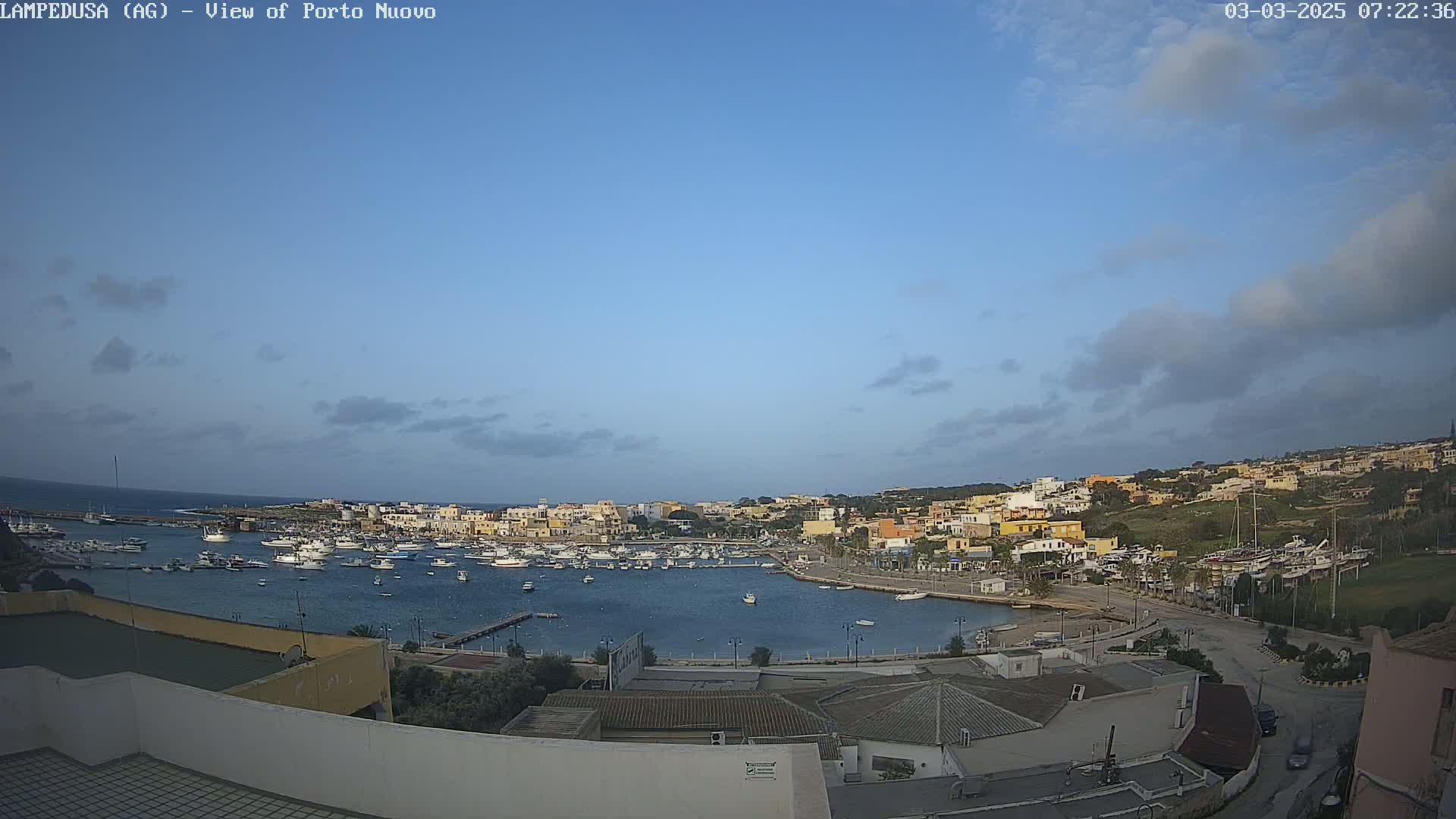 A harbor filled with boats is nestled in a town of low-rise buildings under a mostly clear, light blue sky with a few clouds.