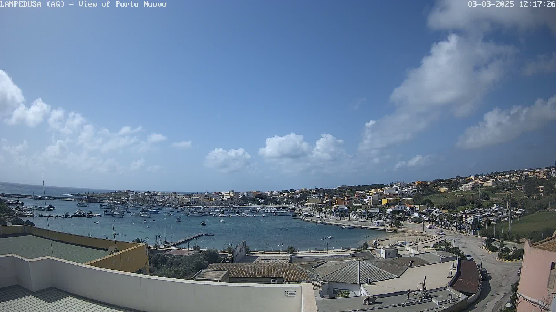 A harbor filled with boats is nestled in a town situated on a hill overlooking the ocean under a mostly sunny sky with scattered clouds.