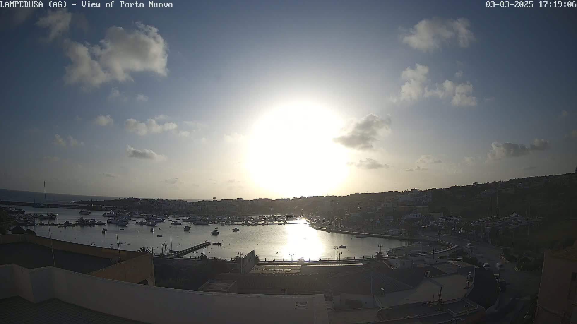 A mostly sunny day with some scattered clouds overlooks a harbor filled with boats and a town built on a hillside.