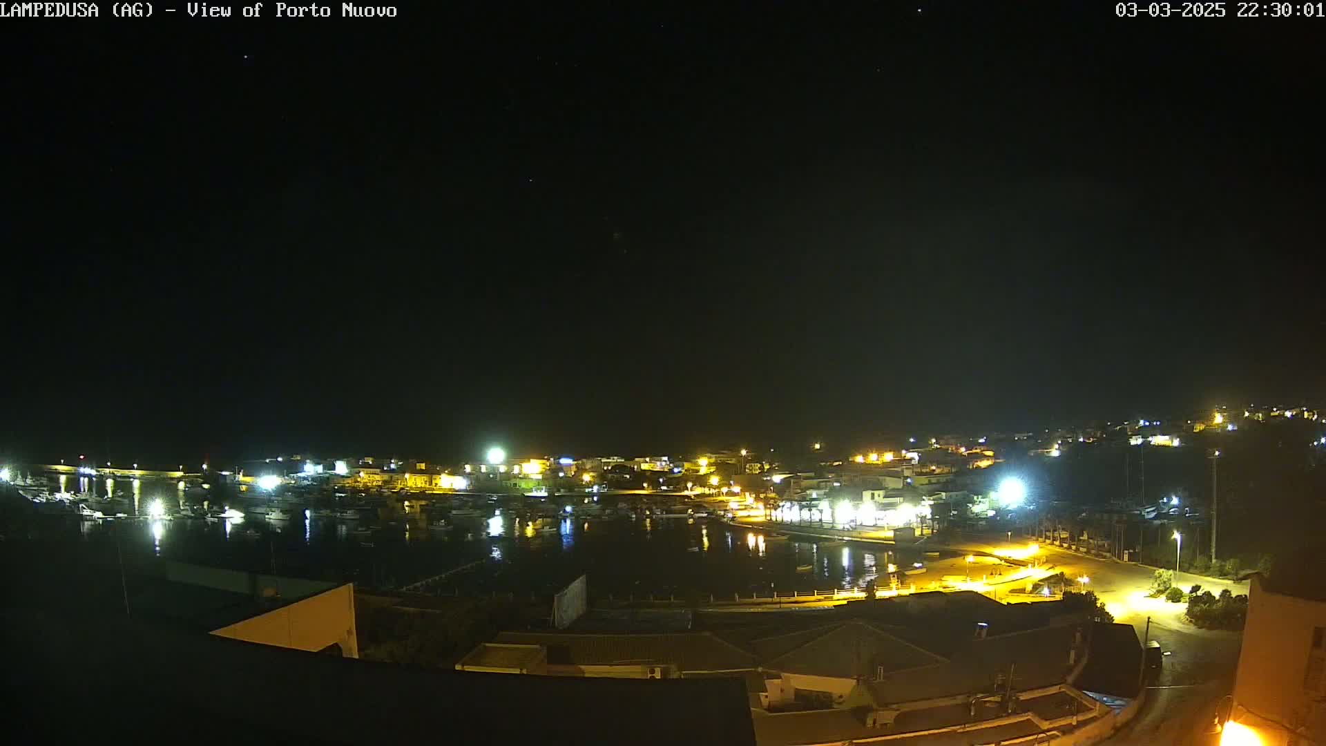 A nighttime view of a harbor town with boats in the water, illuminated by streetlights under a clear, dark sky.