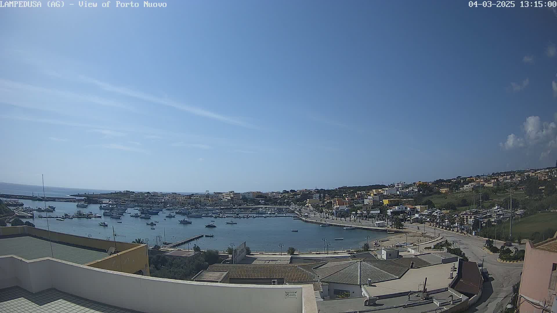 An aerial view of a harbor filled with boats, next to a town under a mostly clear blue sky.