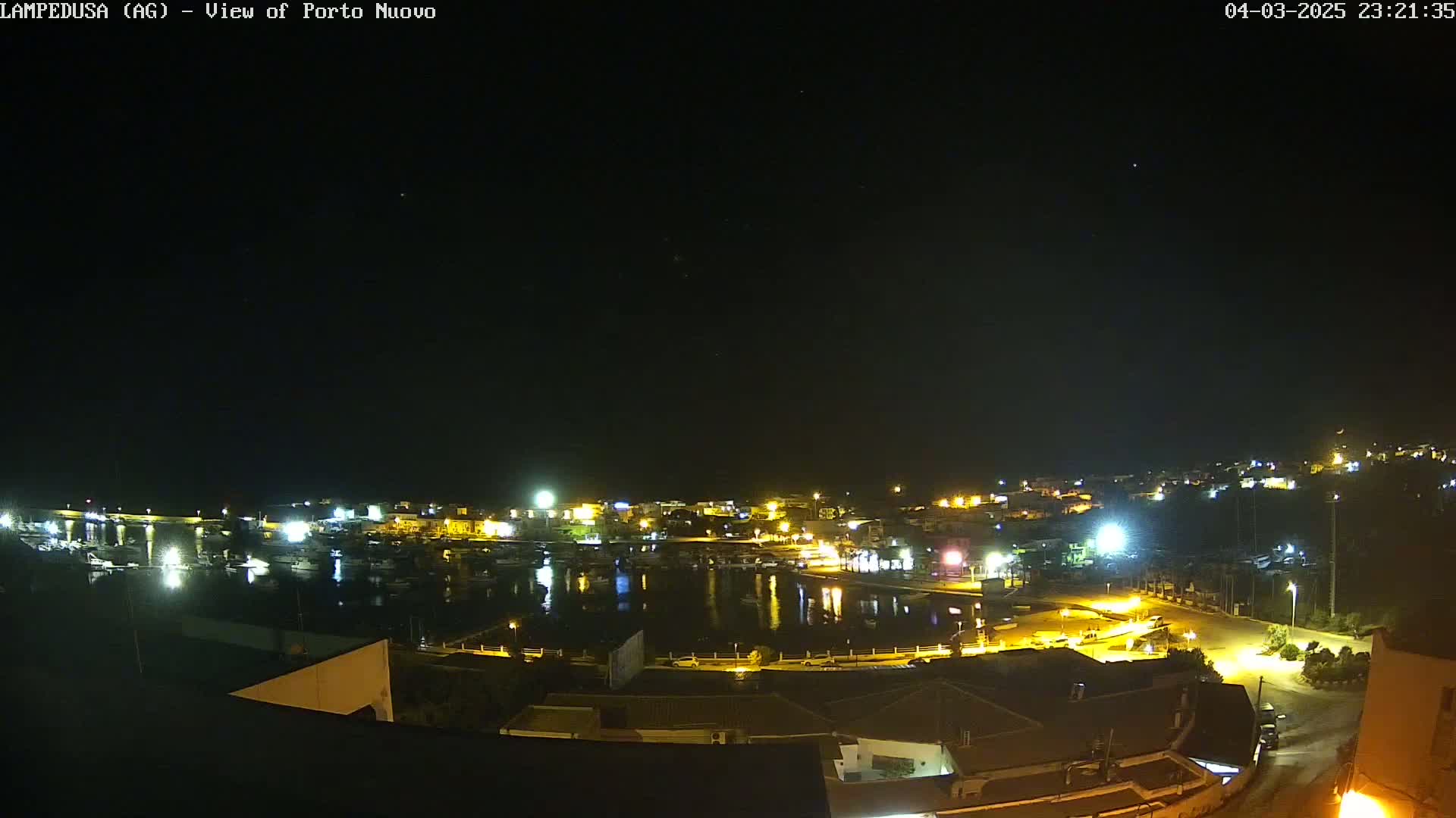 A nighttime, aerial view of a harbor town with its lights illuminating the water and buildings under a clear, starry sky.