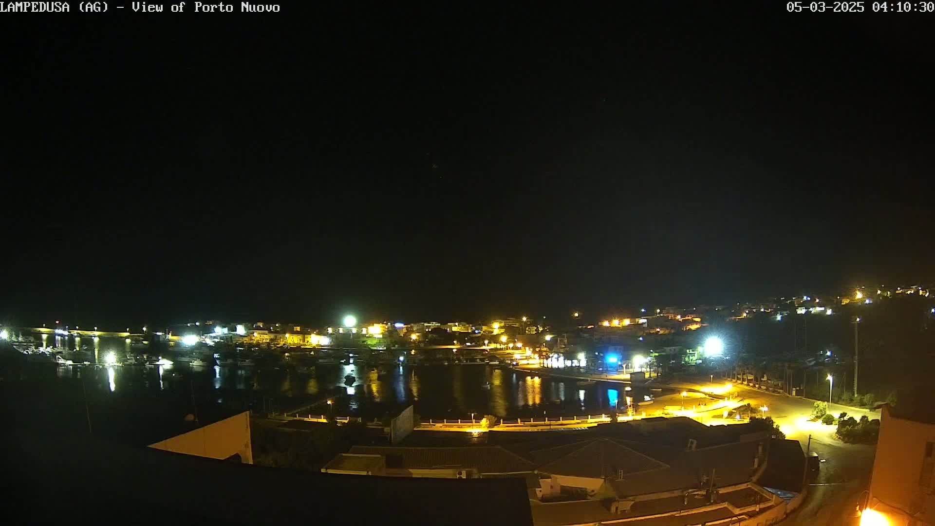 A nighttime, high-angle view of a harbor town with boats in the water and buildings lit up along the shoreline under a clear, dark sky.
