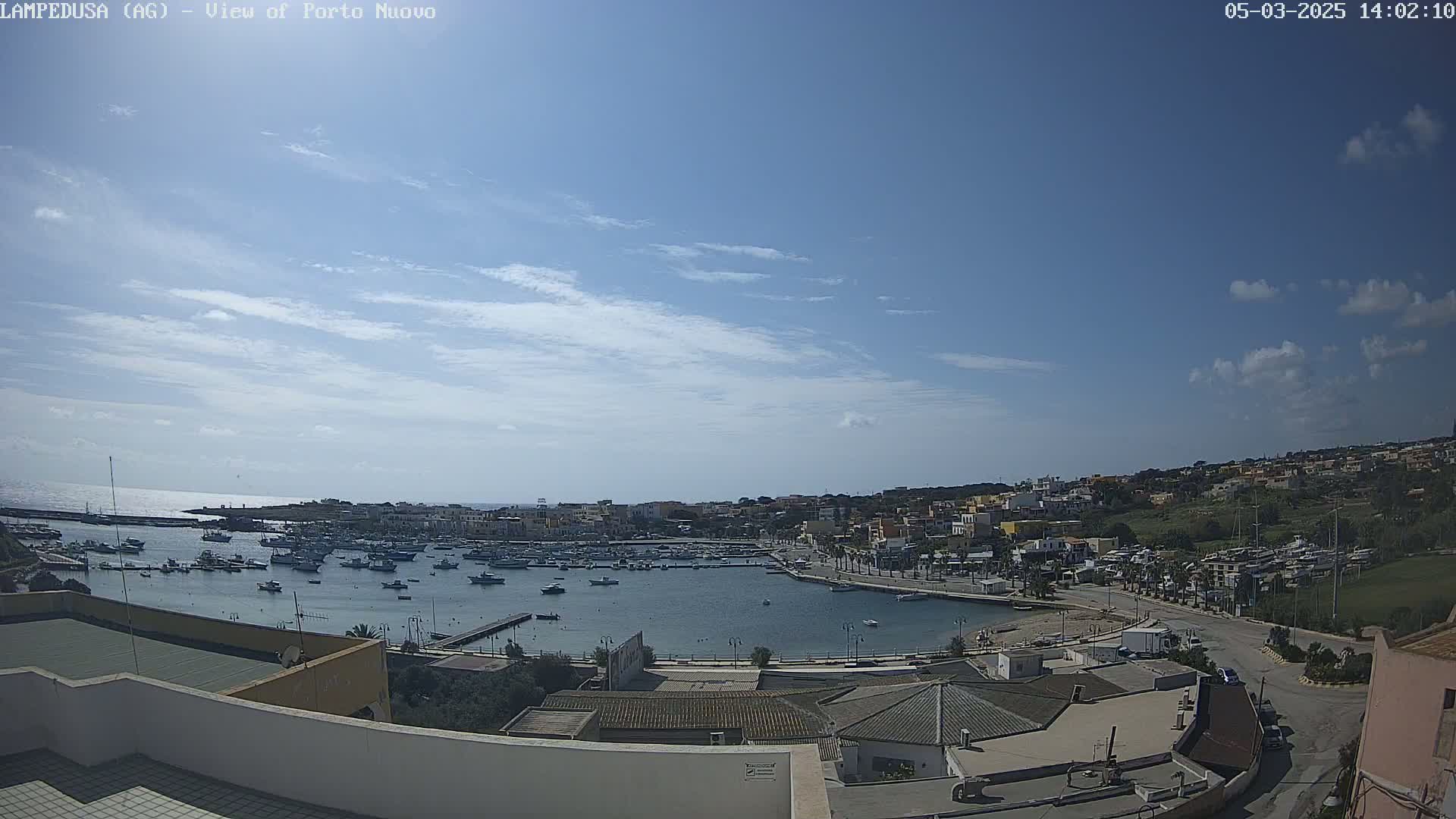 A harbor filled with boats is situated below a mostly clear blue sky with scattered clouds, overlooking a town built on a hillside.