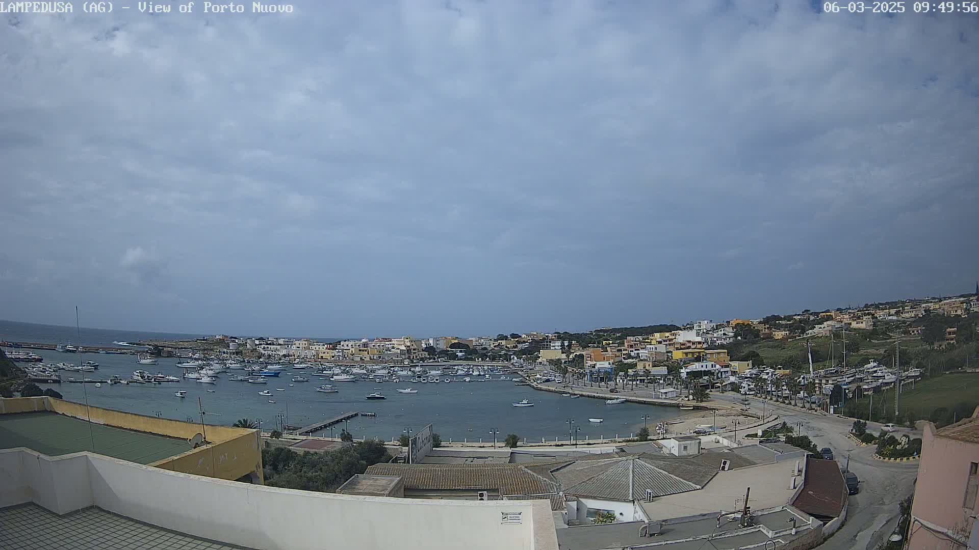 A harbor filled with boats is nestled in a town of low-rise buildings under a mostly cloudy sky.