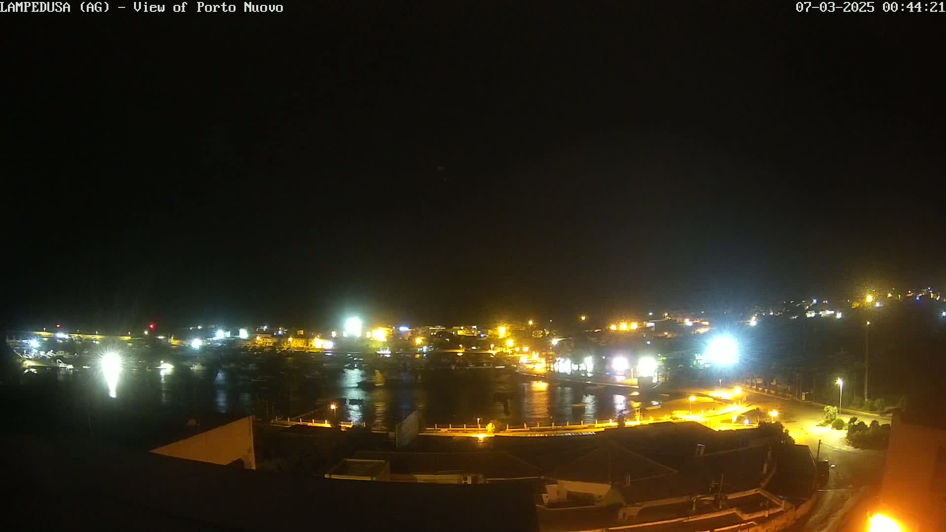 A nighttime aerial view shows a harbor town illuminated with many lights, under a clear, dark sky.