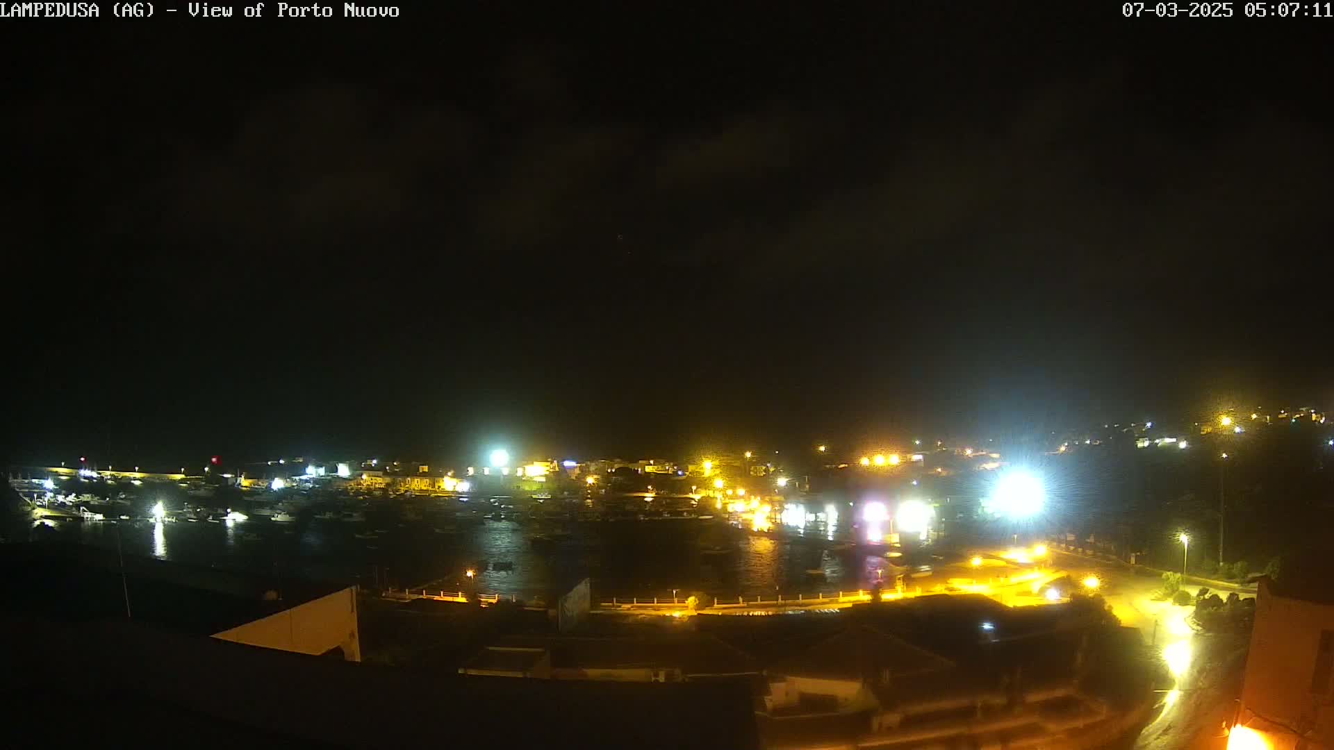 A nighttime, overhead view of a harbor filled with boats, illuminated by the lights of a town on a seemingly clear night.