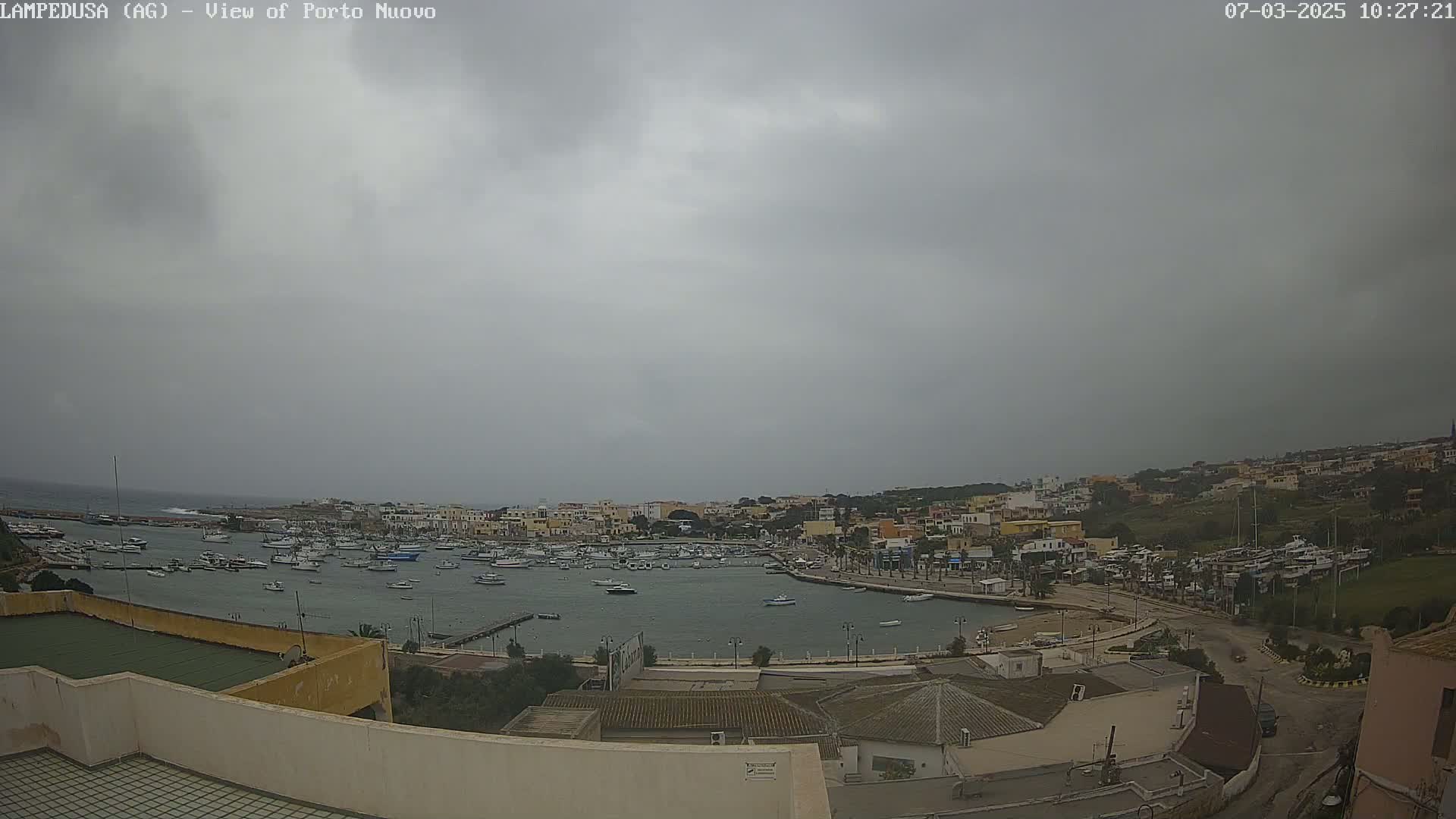 A harbor filled with boats is visible under an overcast sky, with a town and surrounding landscape in the background.