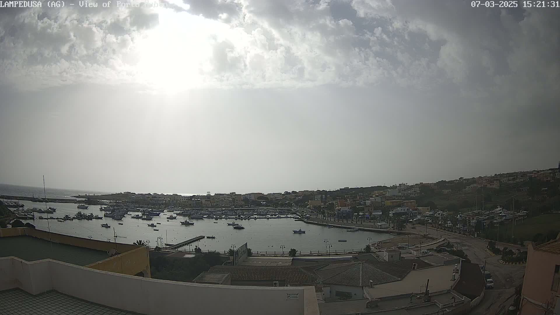 A harbor filled with numerous boats is visible beneath a partly cloudy sky, with the sun somewhat obscured.