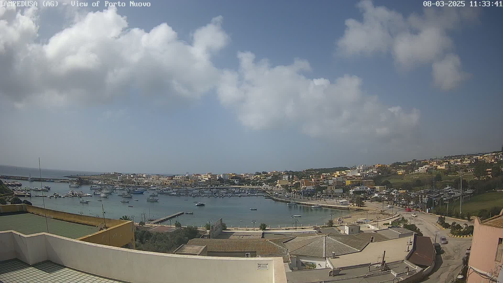 A partly cloudy sunny day overlooks a harbor filled with boats and a town of low buildings on a hillside.
