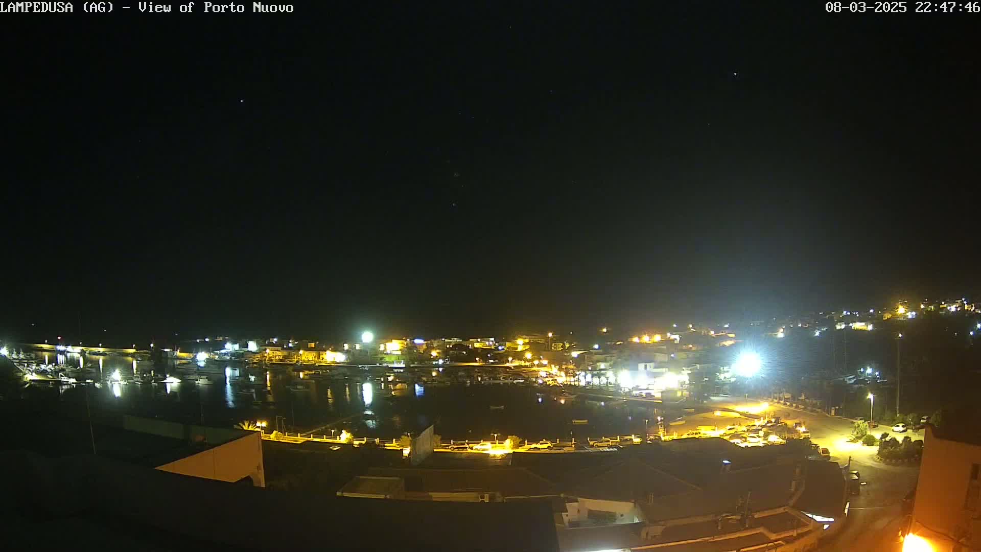 A nighttime, high-angle view of a harbor town with many lights illuminating boats in the water and buildings on the shore under a clear, starry sky.