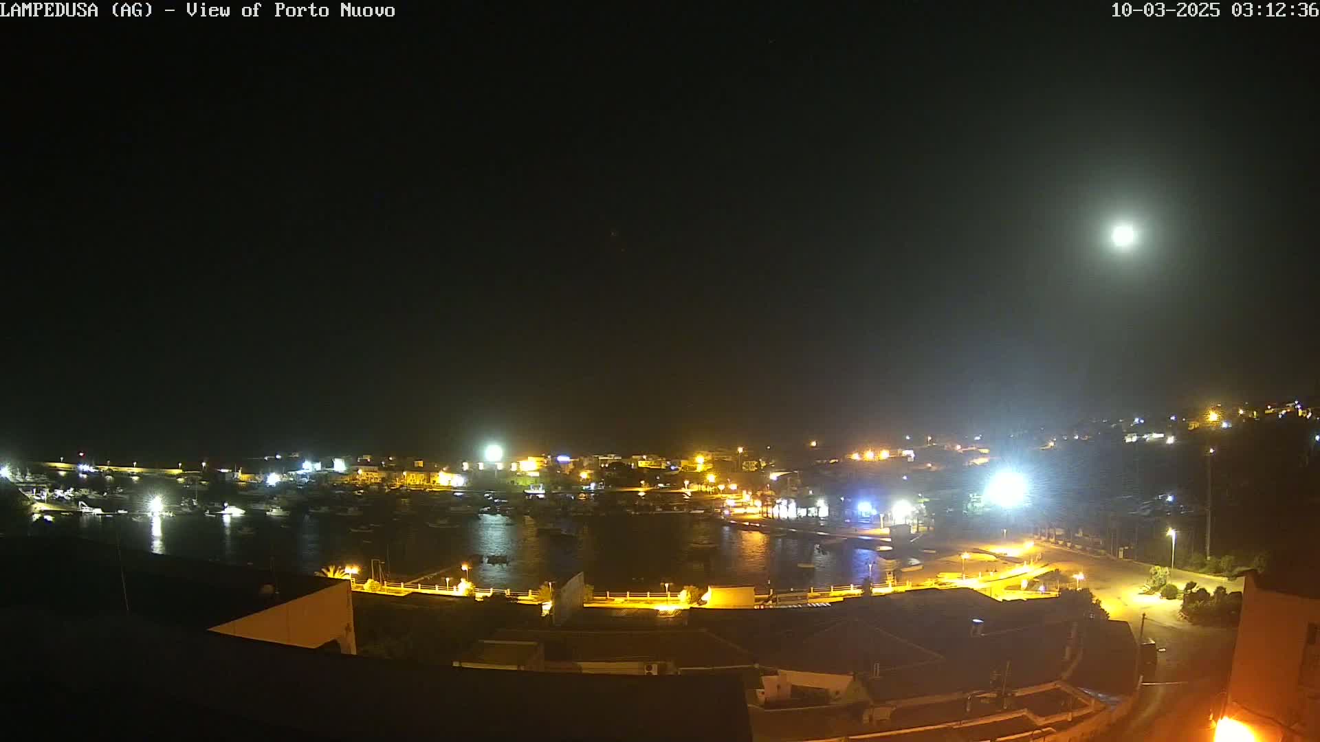 A nighttime, brightly lit harbor with boats and buildings under a full moon.