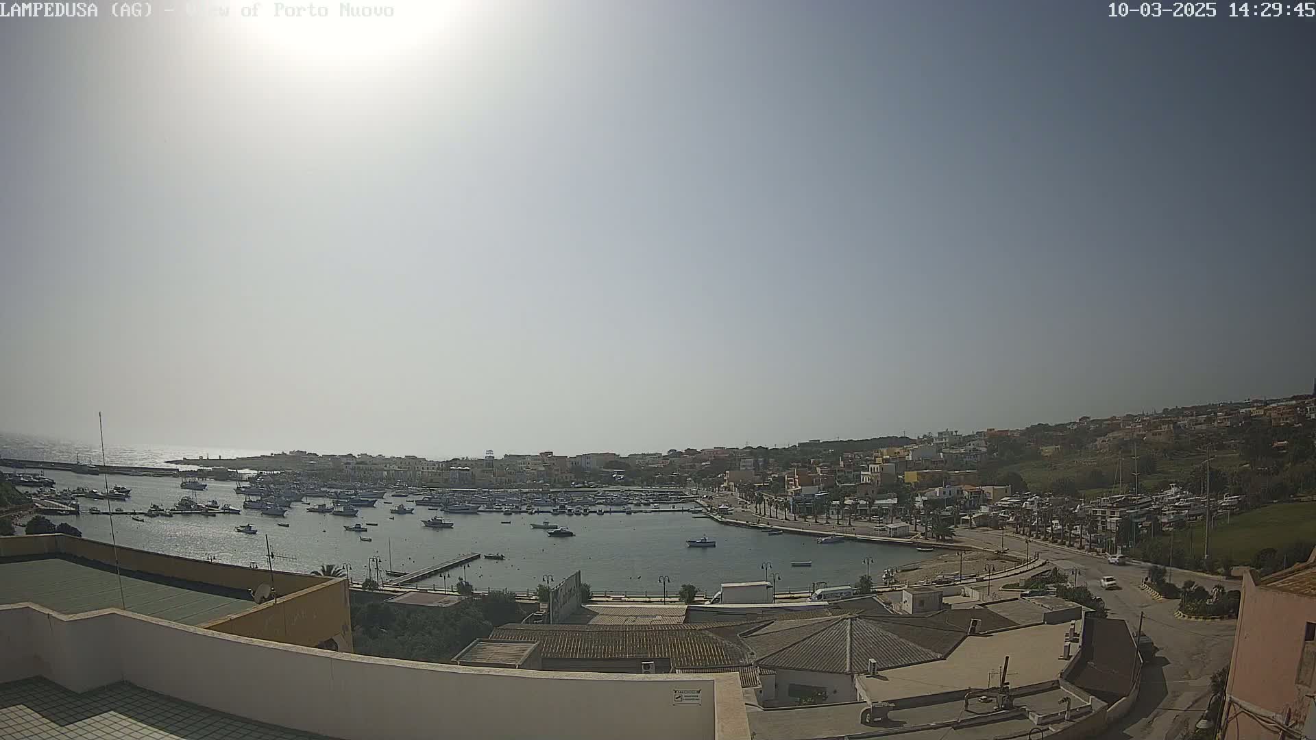 An aerial view shows a harbor filled with boats, a town on a hillside, and a bright, sunny sky.