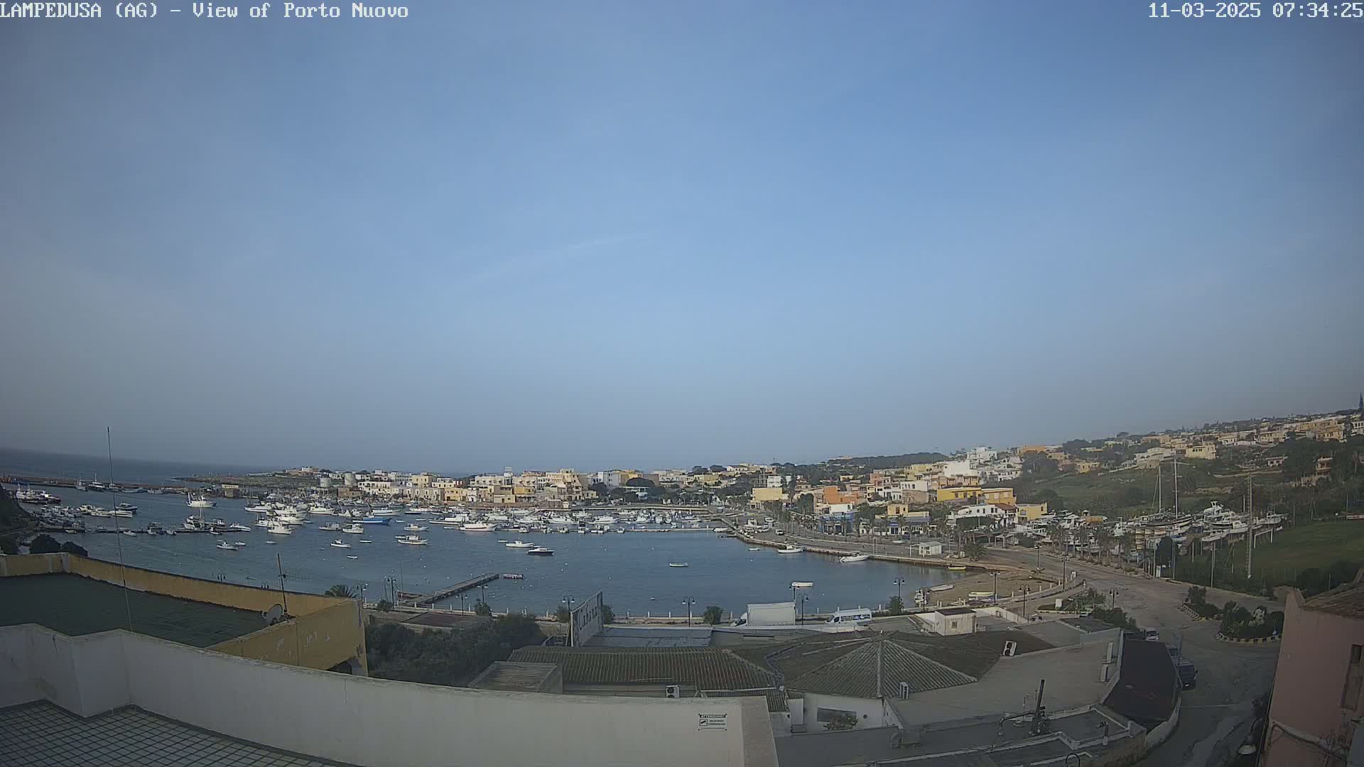 A harbor filled with boats is nestled in a town of low-rise buildings under a clear, light-blue sky.