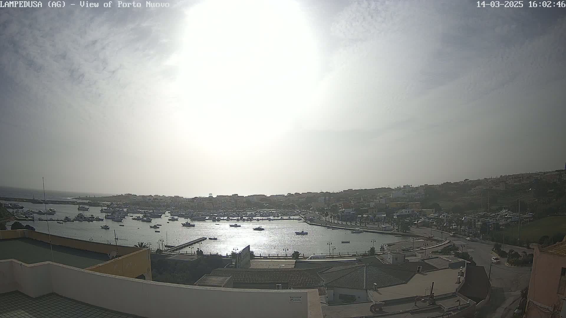 A hazy sun shines over a harbor filled with numerous boats and a town built into a hillside.