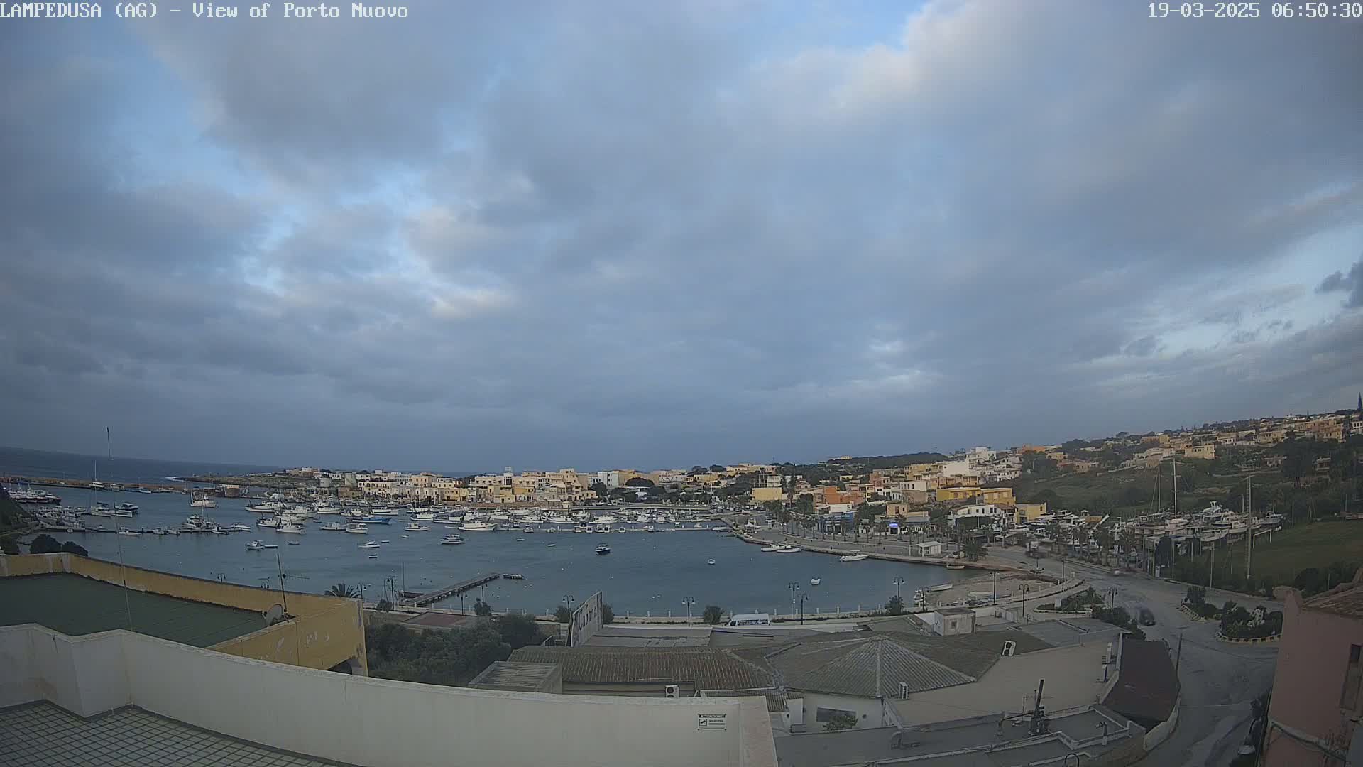 A harbor filled with boats is nestled within a town of low-rise buildings under a mostly cloudy sky.