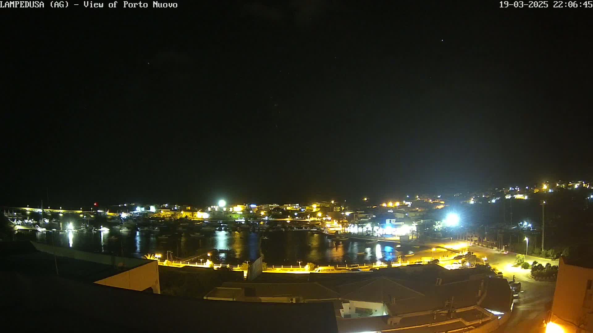 A nighttime view of a harbor town with boats in the water, illuminated by artificial lights under a clear, starry sky.