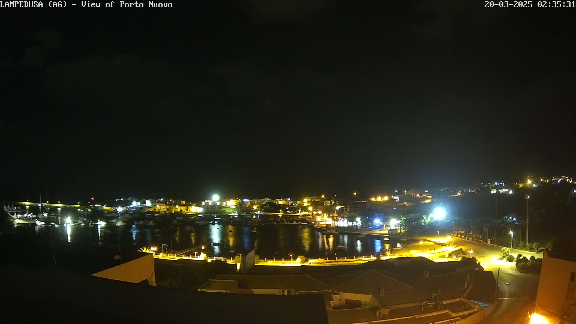 A nighttime, high-angle view of a harbor town with boats in the water and illuminated buildings along the shore under a clear, dark sky.