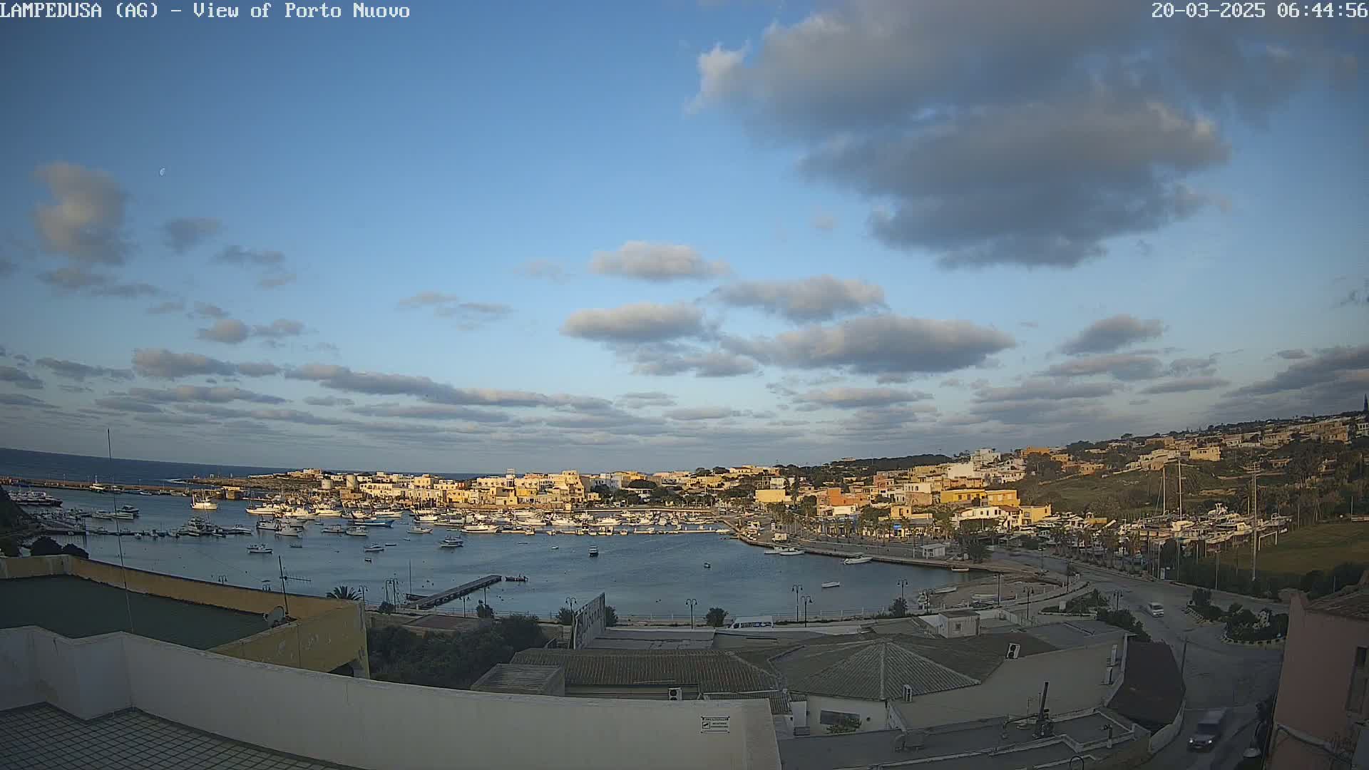 A partly cloudy sky overlooks a harbor filled with boats and a town of low-rise buildings.