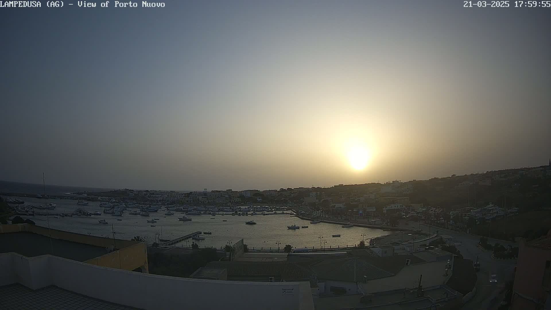 A hazy sunset illuminates a harbor filled with boats, and a town nestled on a hillside.