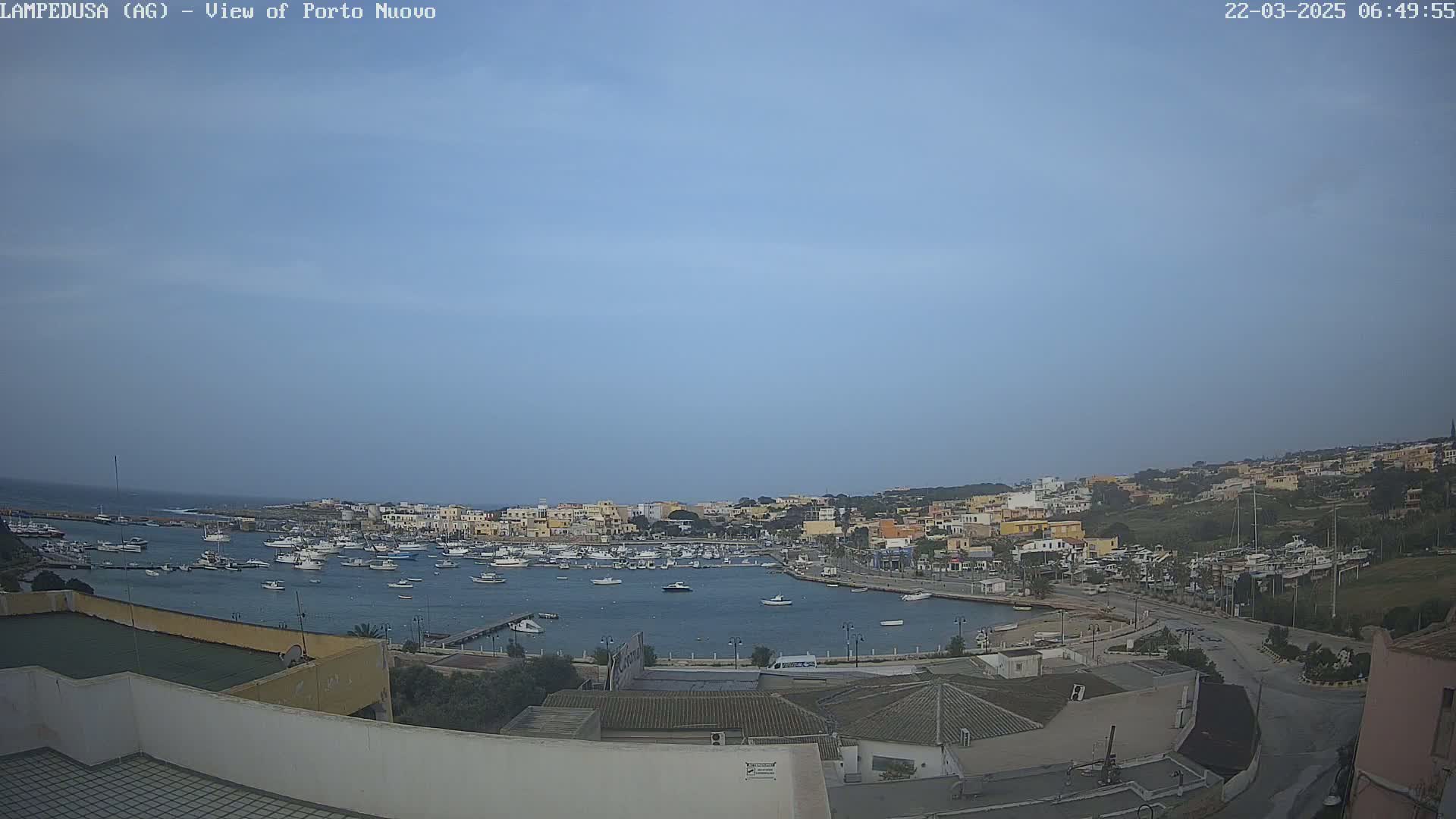 A harbor filled with boats is nestled in a town of low-rise buildings under a mostly clear, light-blue sky.