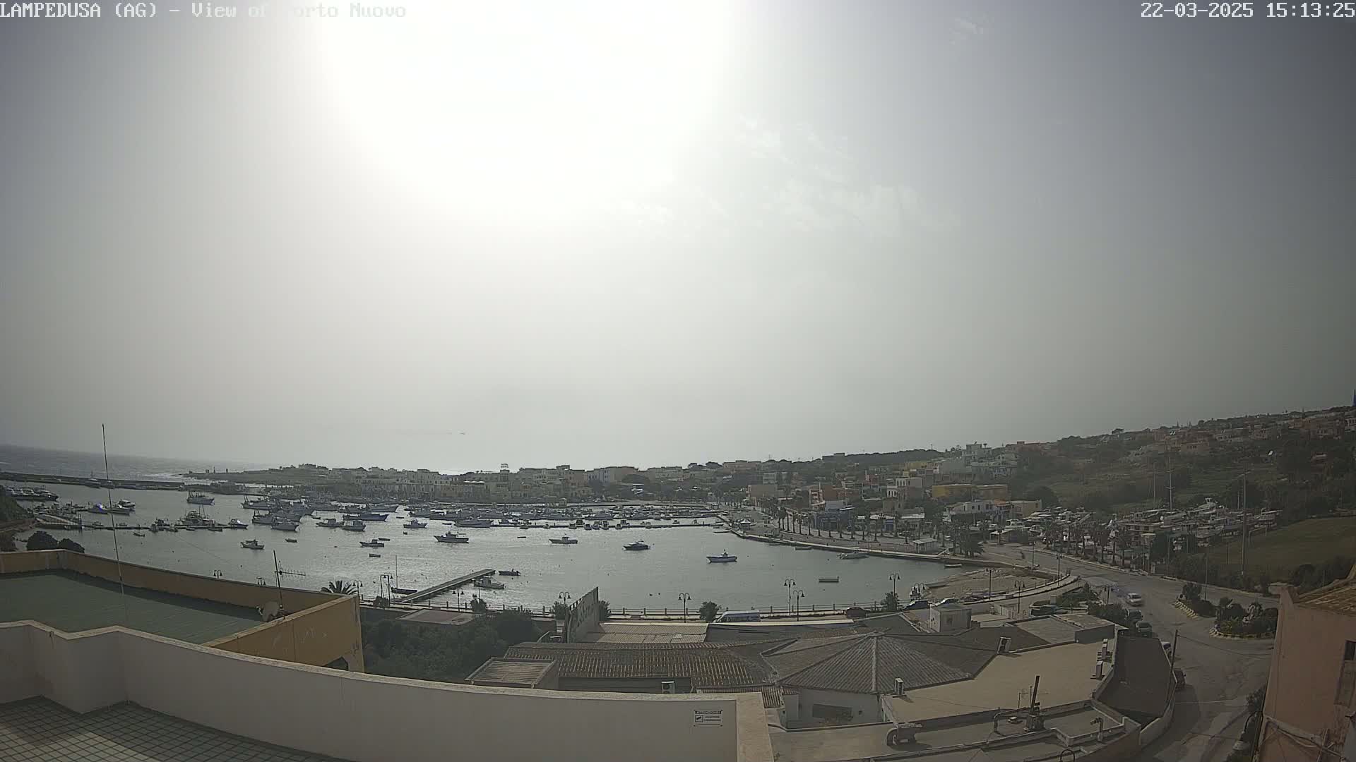 A hazy, sunny day overlooks a harbor filled with numerous boats and a town situated on a hillside.