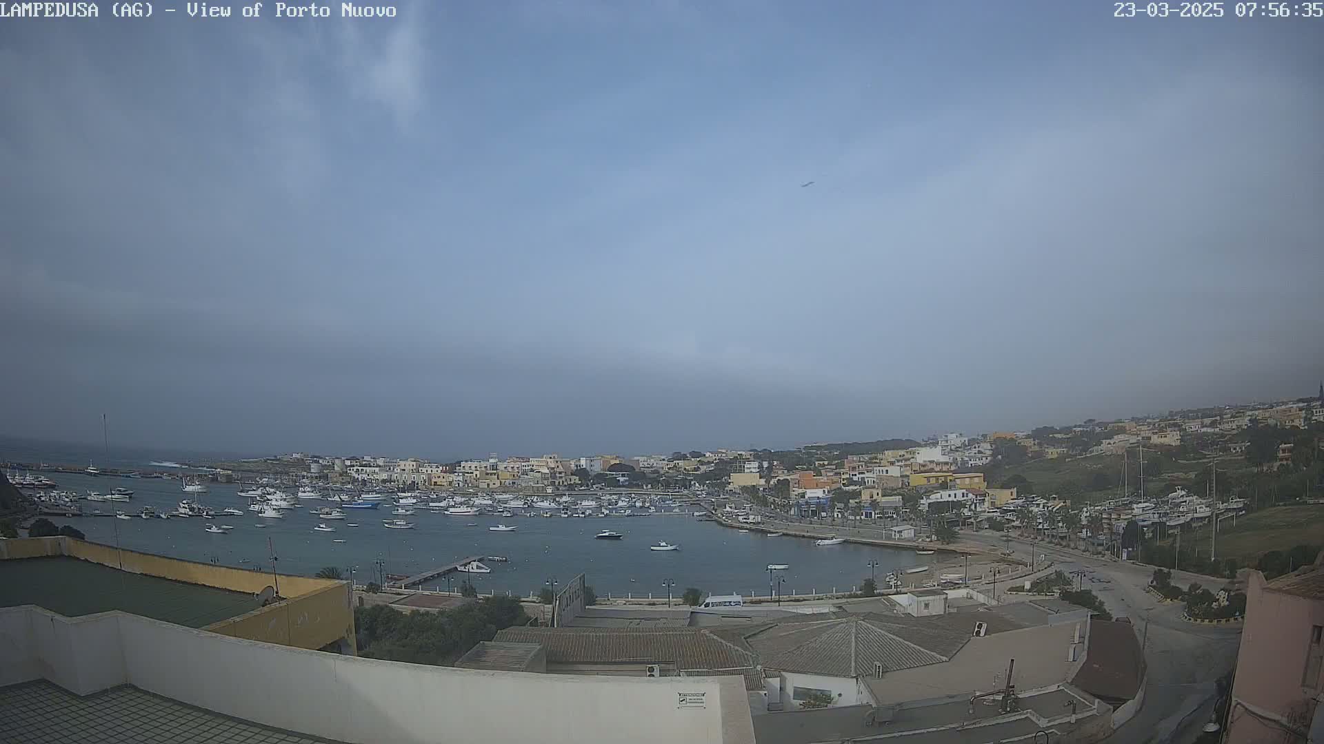 An overcast sky overlooks a harbor filled with boats and a town of low buildings on a hilly coastline.