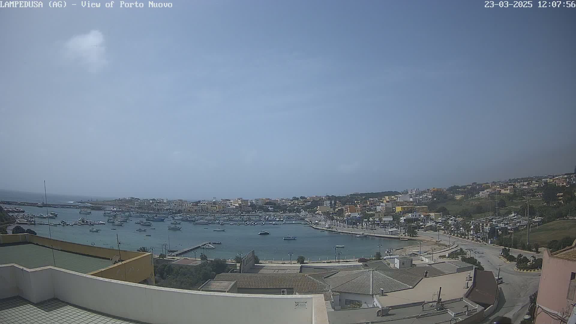 A mostly sunny day reveals a harbor filled with boats, surrounded by a town built on a hillside.