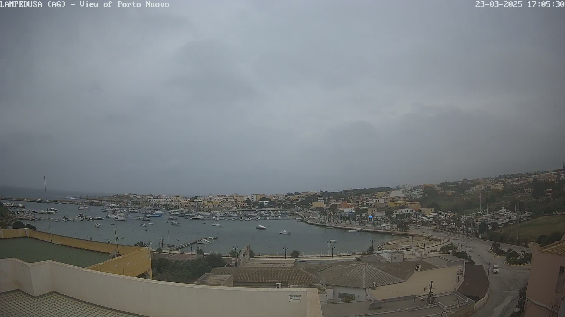An overcast sky hangs over a harbor filled with numerous boats, nestled within a town of low-rise buildings on a hilly coastline.