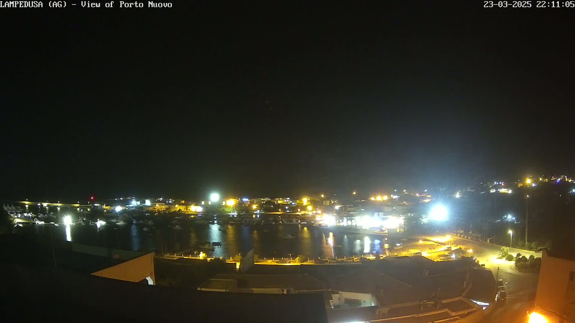 A nighttime view of a harbor town with many boats in the water and lights illuminating the buildings and surrounding area under a clear, dark sky.