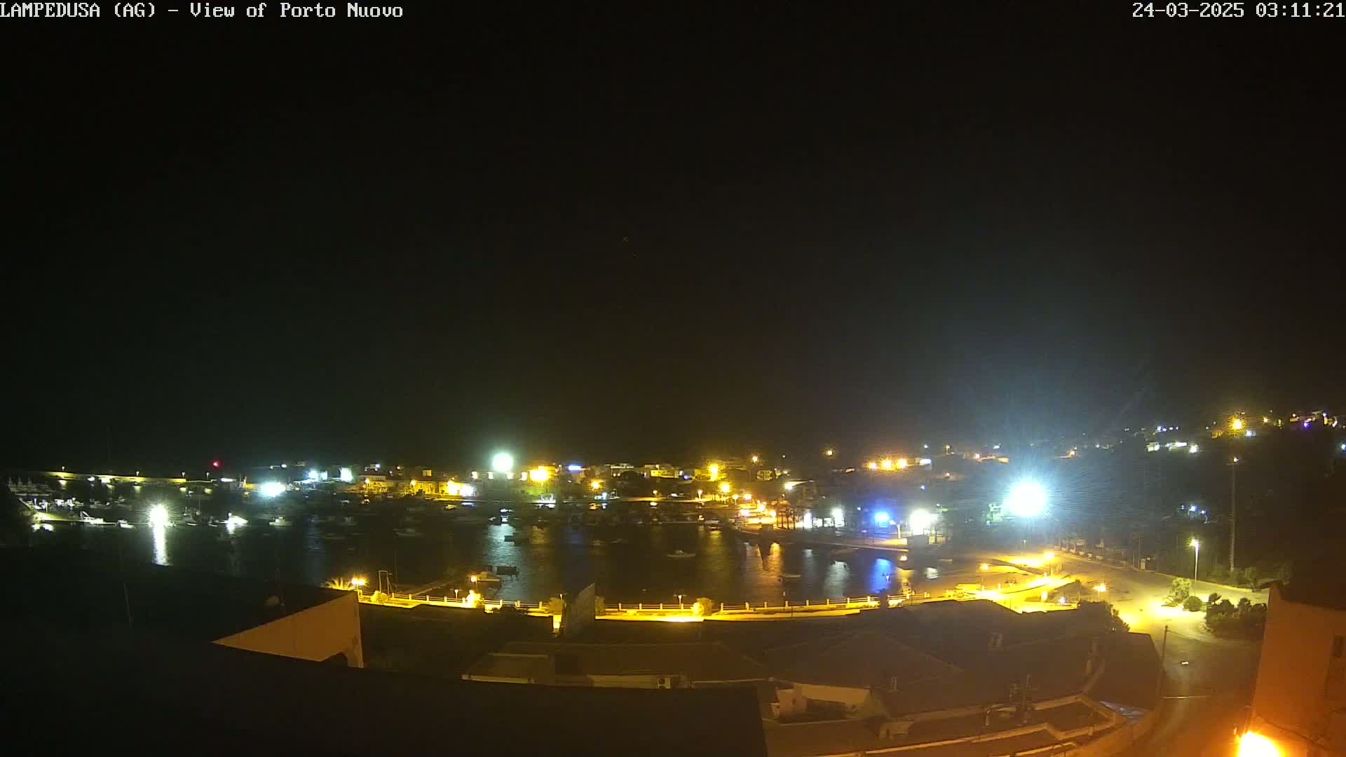 A nighttime, high-angle view shows a harbor filled with boats, surrounded by illuminated buildings and streets.